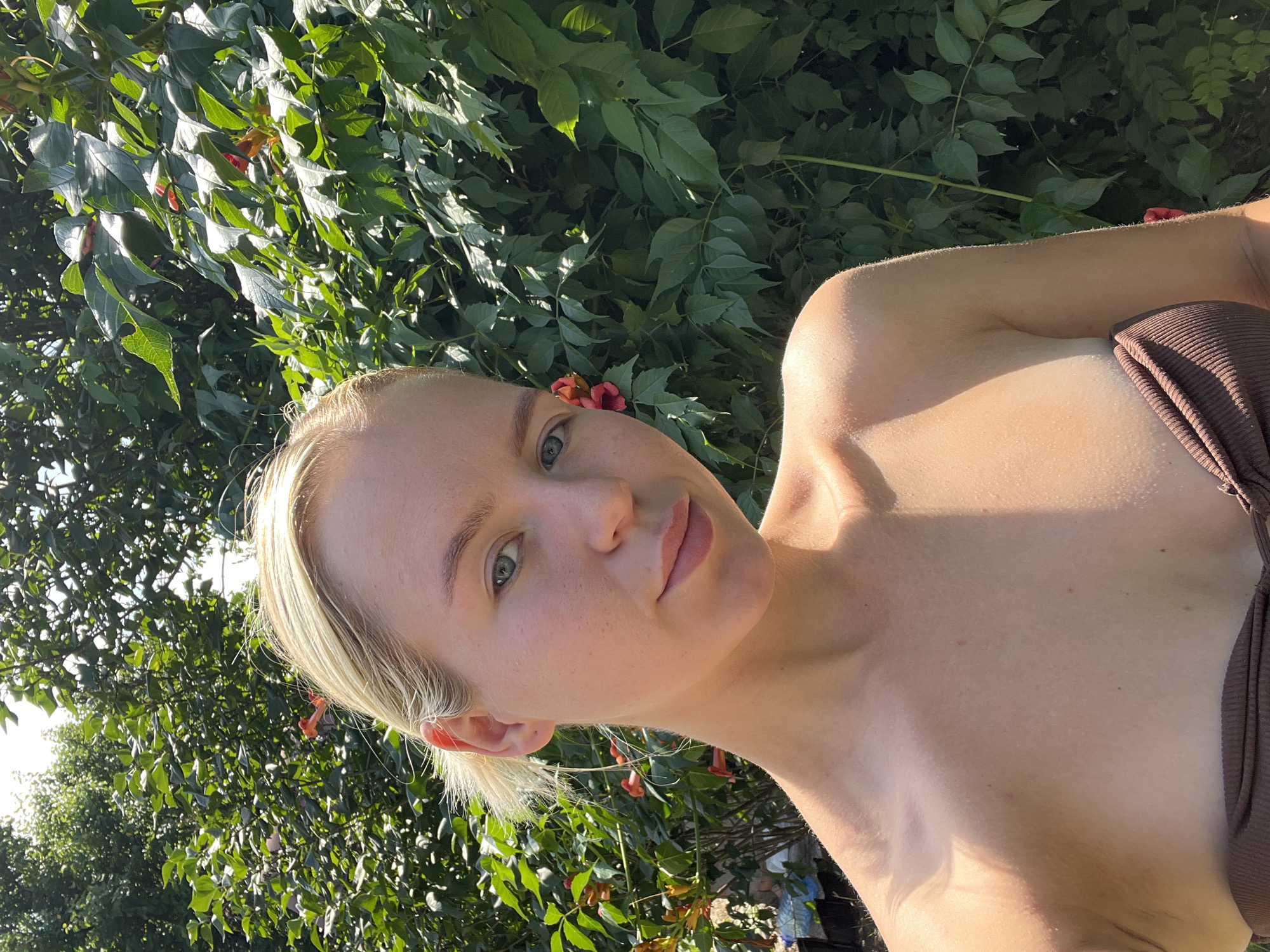 A fair-skinned young woman with blonde hair pulled back is taking a selfie outdoors. She is wearing a brown strapless top and is standing in front of a lush green hedge with some orange flowers. Sunlight is dappling her face and upper body. The background is out of focus.
