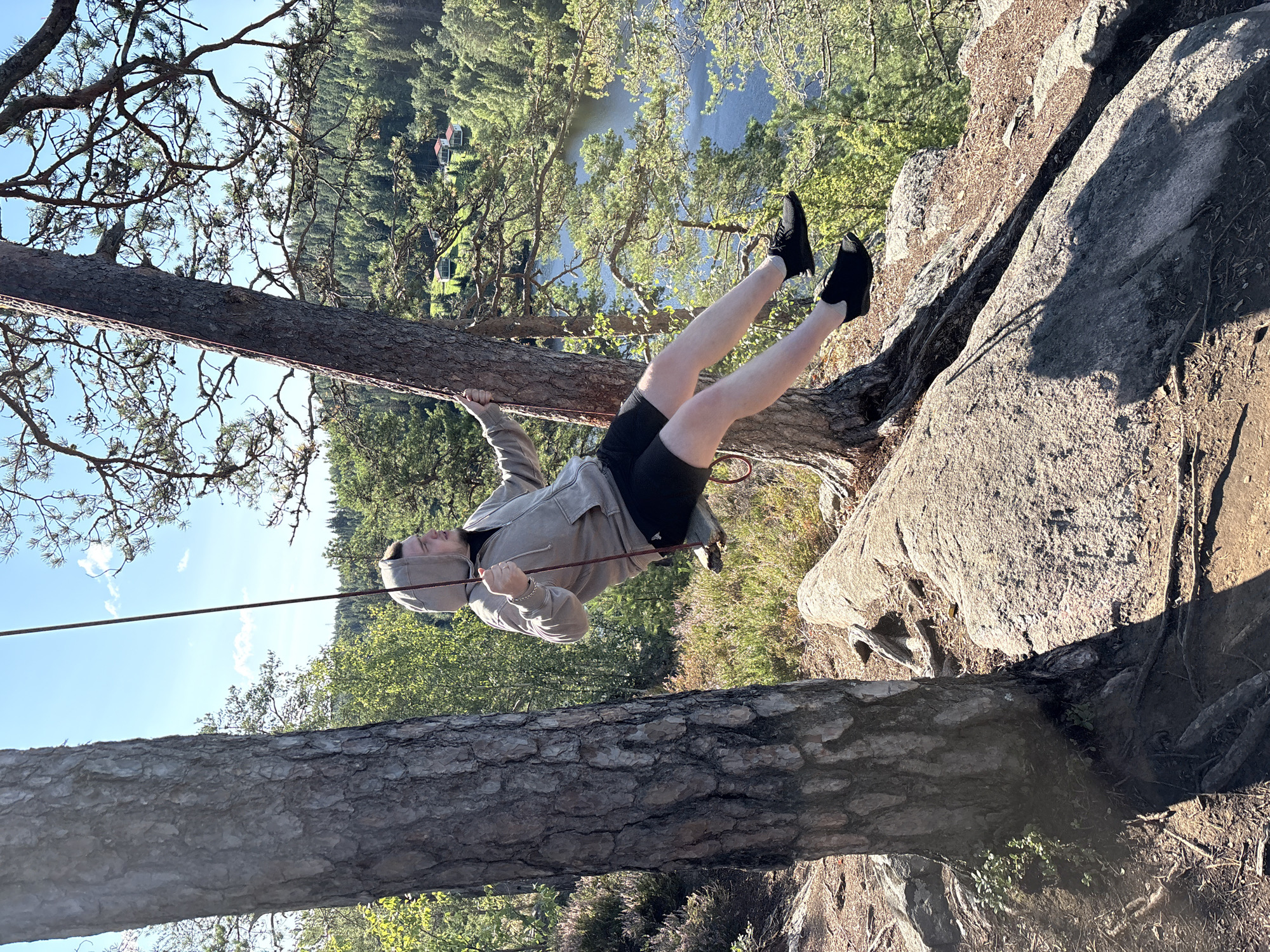 A young man with fair skin and short brown hair is swinging on a homemade swing attached to a tree. He is wearing a gray hooded sweatshirt, black shorts, and black athletic shoes. The swing is made of a wooden plank and a red rope. The man is positioned on the right side of the frame, with his body angled towards the left, his legs extended forward as he swings. His right hand is holding onto the rope.
The background of the image reveals a natural, wooded landscape overlooking a body of water. The area is characterized by a mix of pine trees and deciduous trees, with some undergrowth of heather. The terrain appears rocky and elevated, suggesting a viewpoint. In the distance, across the water, several small houses with red roofs are visible amongst the trees. The sky is blue with scattered white clouds. The lighting indicates it is daytime, with the sun casting shadows on the ground. The specific location is given as Kir'yavalakhti, Russia.