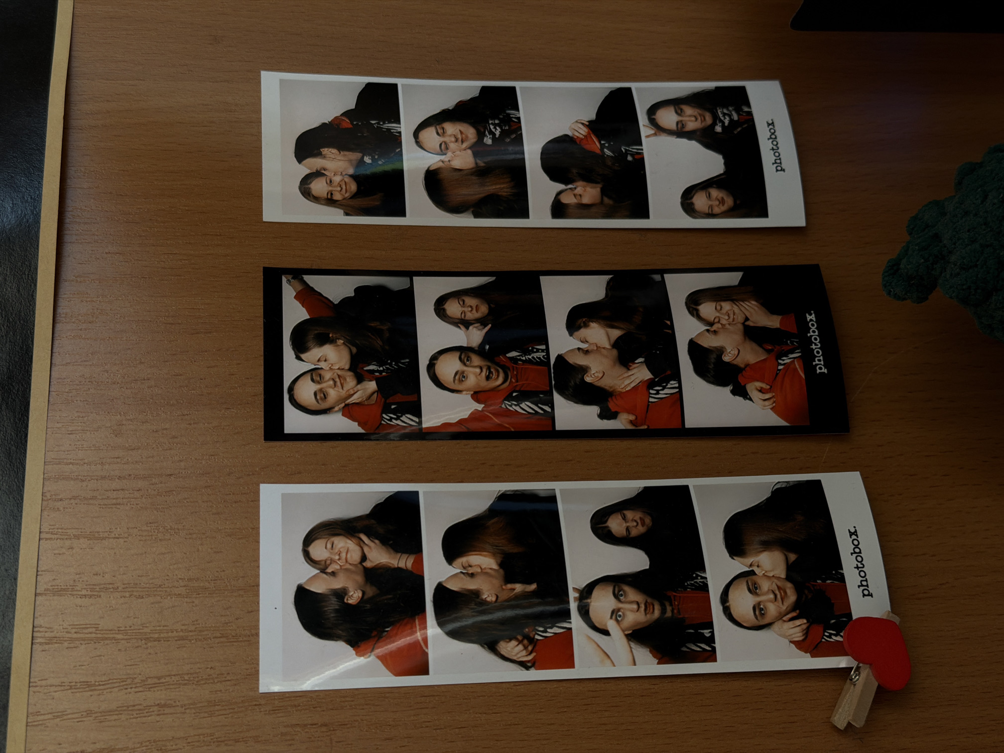 The scene is a wooden surface, likely a tabletop. Three photobooth strips are arranged on the surface. Each strip contains four images of two people, appearing to be kissing or posing affectionately. The photobooth strips are marked with the word "photobox." A small wooden clothespin with a red heart attached is clipped to the bottom right corner of the bottom strip. To the right of the photobooth strips, a portion of a green, crocheted object is visible. There are no indications in the image relating to Perm, Russia.