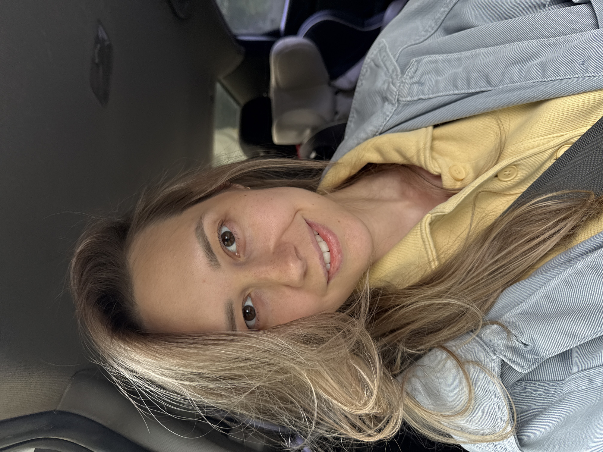 A woman is seated in a car. She is wearing a yellow collared shirt and a light blue denim jacket. The car's interior is dark. In the background, a car seat is visible. The location is likely not Chaikovsky, Russia, as it cannot be determined from the image.