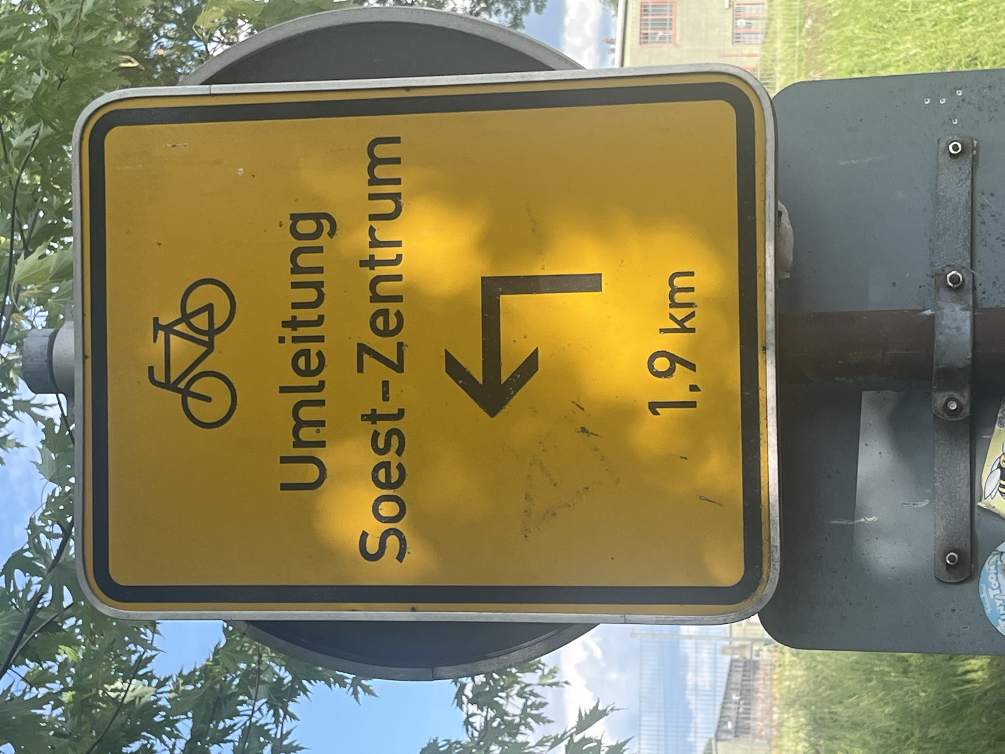 An outdoor, brightly lit image shows a yellow road sign for a bicycle detour, oriented sideways. The rectangular sign features a prominent black bicycle symbol in the upper left corner. Below it, large black text reads "Umleitung Soest-Zentrum," indicating a detour to the town center of Soest. An arrow points downwards and then turns sharply to the right, signifying the direction of the detour. In the lower right corner, the distance "1,9 km" is displayed.
The sign has a black border and is mounted on a dark grey, somewhat weathered metal pole or frame. Green foliage from a tree is visible in the upper and lower left portions of the frame, suggesting a natural setting. The background includes a clear blue sky with some white clouds, a light-colored building with multiple rectangular windows, a fence, and green grass, all bathed in bright sunlight. The overall impression is of a sunny day. The metal frame holding the sign shows some wear and tear, and a small, round sticker featuring a bee is partially visible on the far right.