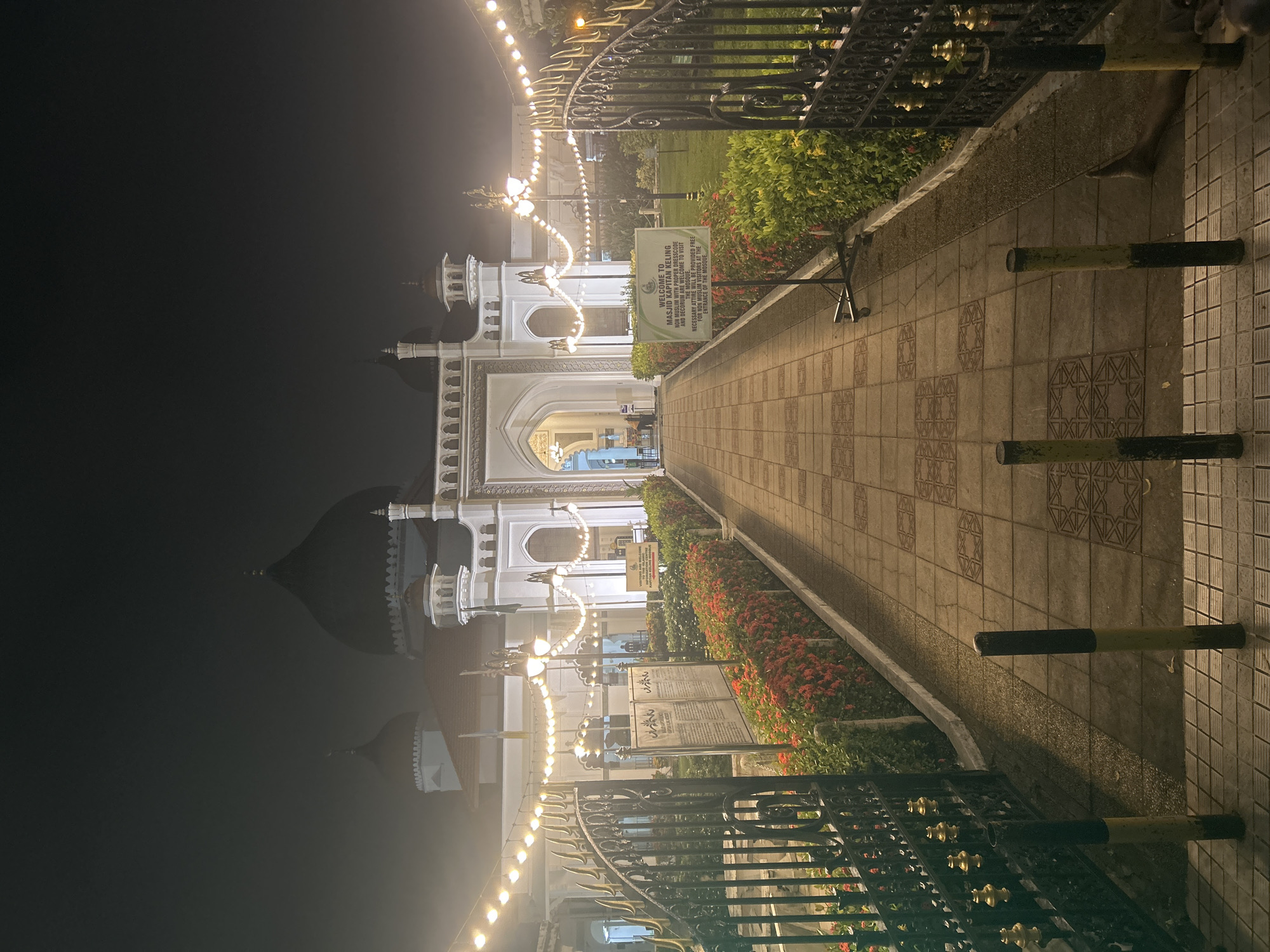The image shows the Kapitan Keling Mosque in George Town, Malaysia at night. A pathway paved with patterned tiles leads through an open gate towards the mosque's entrance. The path is flanked by neatly trimmed hedges of red and green foliage. String lights are hung across the entrance area, illuminating the white-tiled facade of the mosque, which features domes and minarets. Two informational signs are visible on the left and right sides of the pathway, with the sign on the right displaying a welcome message for visitors. The mosque is bathed in a soft, ambient light, creating a serene atmosphere. The sky is dark and hazy, suggesting a foggy or misty condition. Several bollards are placed along the pathway, and the ground shows signs of wetness. A person's leg is visible on the lower right side of the frame, indicating their presence at the scene.