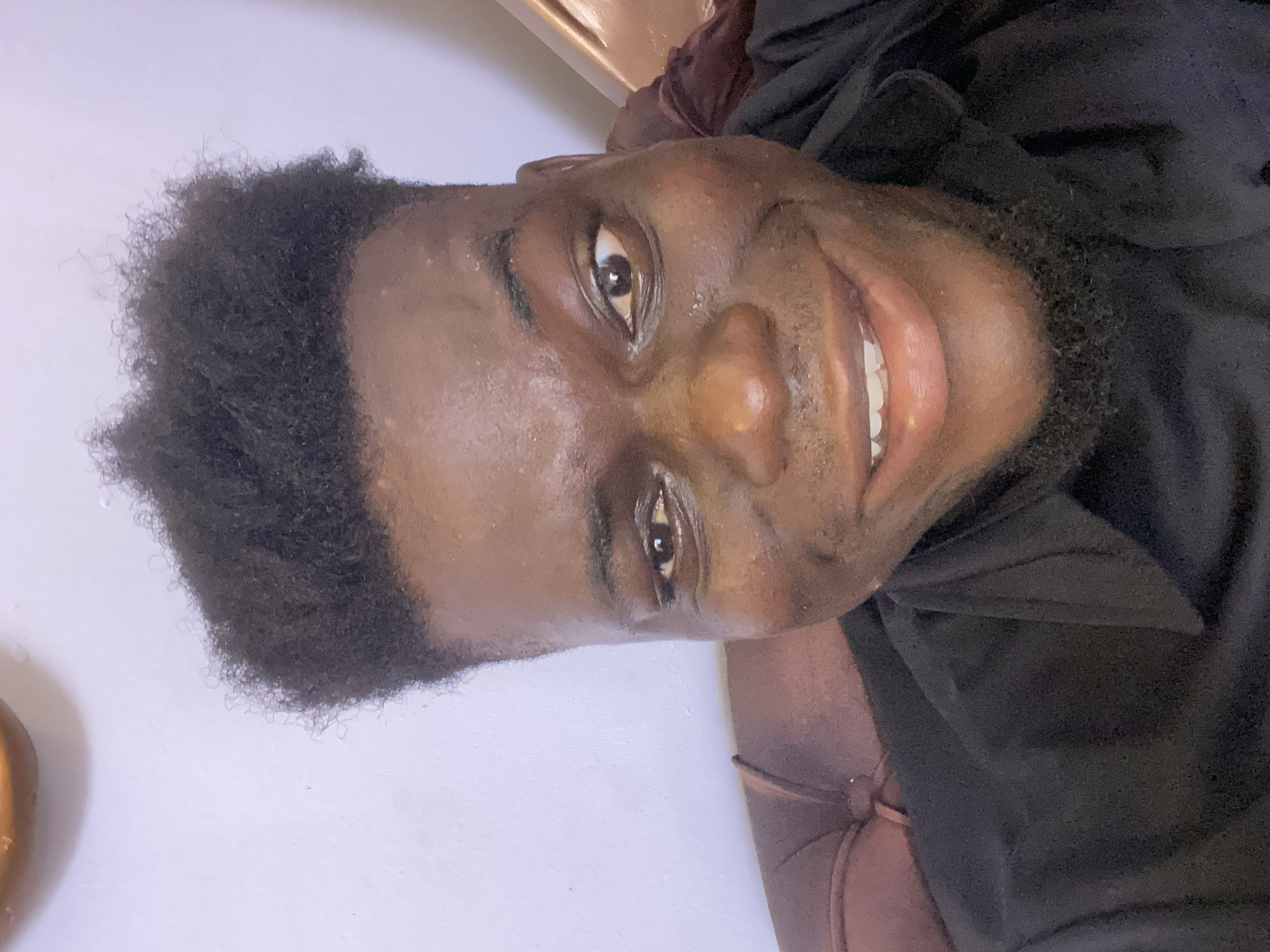 The image shows a man with dark skin, short black hair, and a beard smiling at the camera. He appears to be wearing a black top and is sitting in a brown chair. The background is a light wall. There is no indication of the location in the city of Unknown, Unknown.