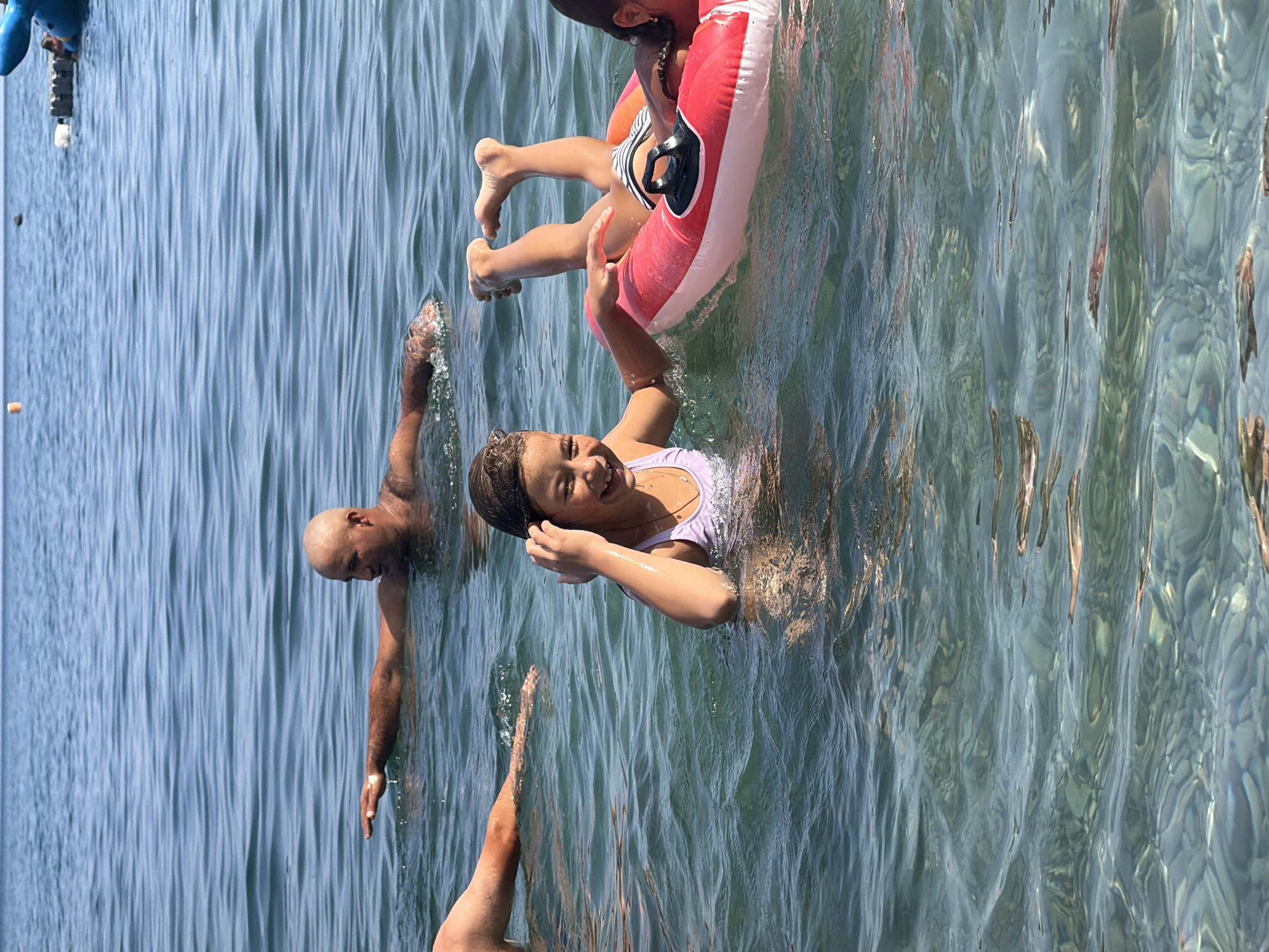 In the city of Partenit, Ukraine, a group of people are enjoying a day at the beach. The water is clear and shallow, with sunlight glinting off the surface. In the foreground, a young girl with dark hair is smiling and looking at the camera, her hand touching her ear. She is wearing a purple swimsuit. Beside her, another young girl with dark hair is lying on a pink and white inflatable tube, her feet pointing upwards. In the background, a bald man with dark skin is swimming with his arm extended. The scene depicts a relaxed and joyful atmosphere at the seaside.