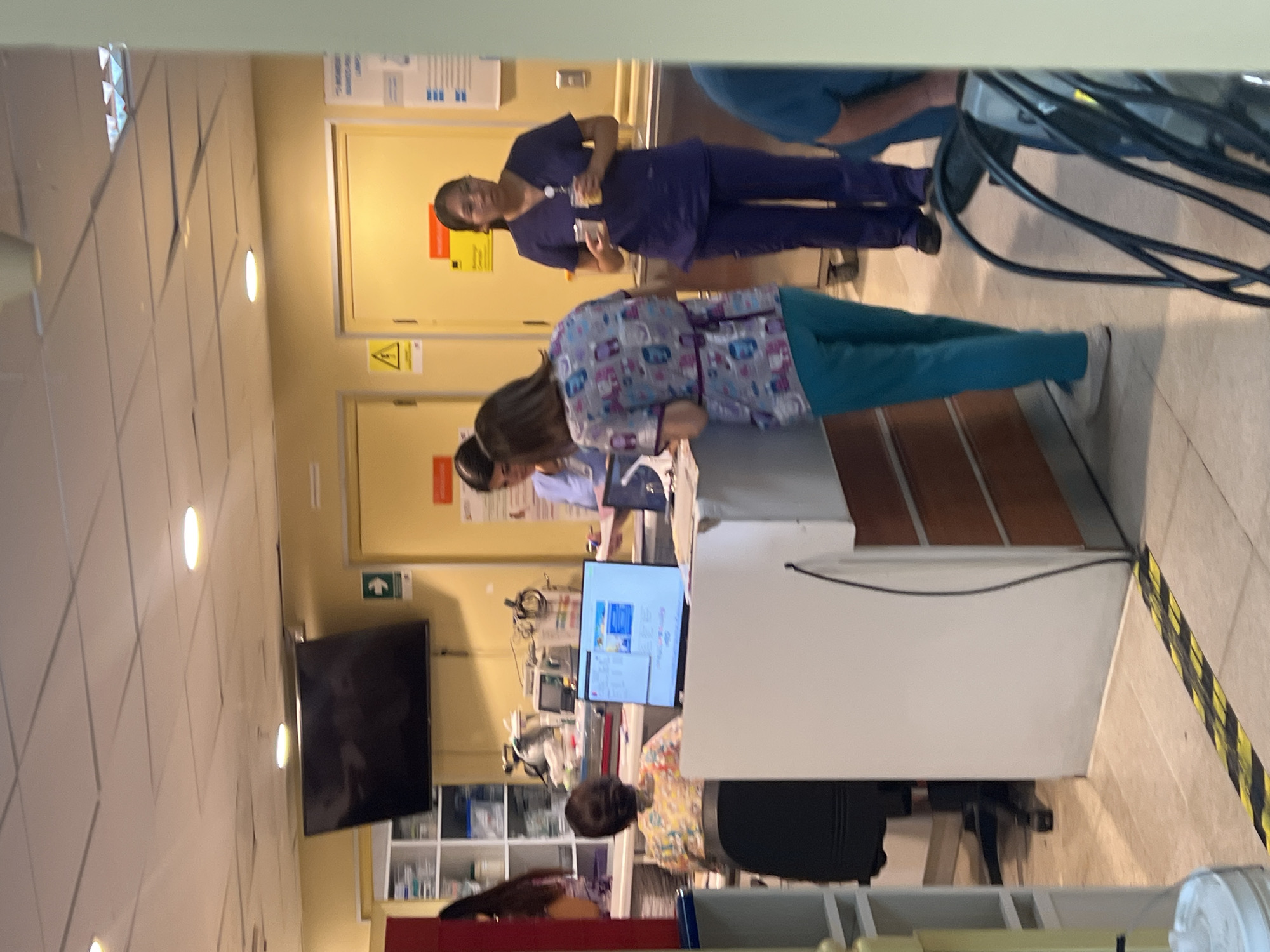 The image depicts a medical environment, likely a hospital or clinic, potentially located in Arica, Chile, based on the general visual characteristics. Multiple individuals, presumably medical staff, are present. Several are wearing scrubs in various colors including dark blue and patterned blue and white tops with teal pants. Some appear to be looking at a computer monitor on a white desk or counter. Medical equipment is visible in the background, including monitors, shelves containing supplies, and other devices. A large screen television is mounted on the wall. The lighting is bright, and the interior design appears functional and sterile.