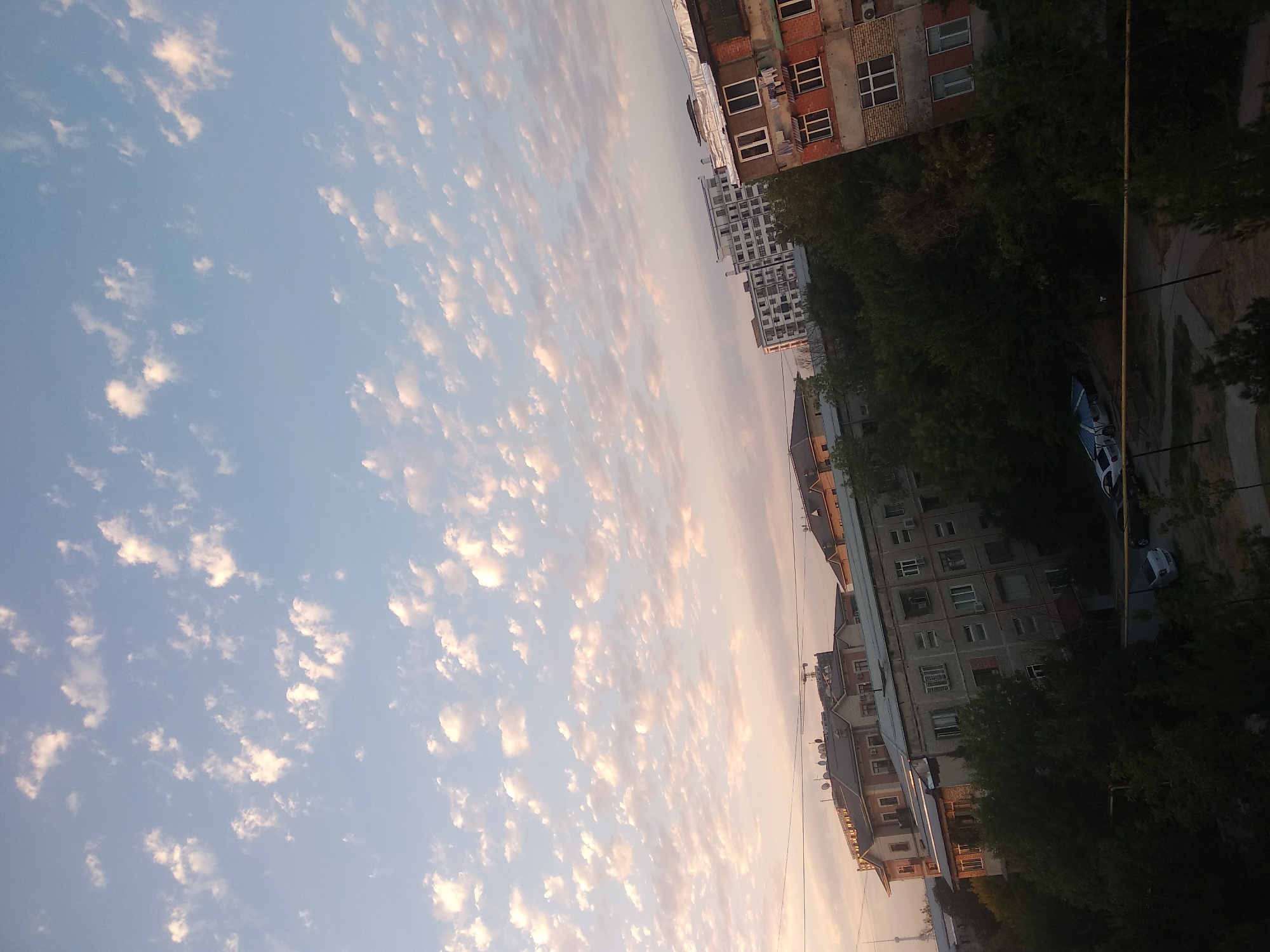 The image depicts an urban landscape in Tashkent, Uzbekistan, under a sky filled with scattered cumulus clouds. The buildings visible include multi-story residential apartments, some with balconies, and a few older structures. Dense green trees are interspersed between and in front of the buildings, suggesting a residential area. In the foreground, a courtyard or street area is visible with parked cars, including a white sedan and a darker vehicle. Laundry is hanging on a balcony of one of the buildings. The sky exhibits a soft light, possibly indicating sunrise or sunset.