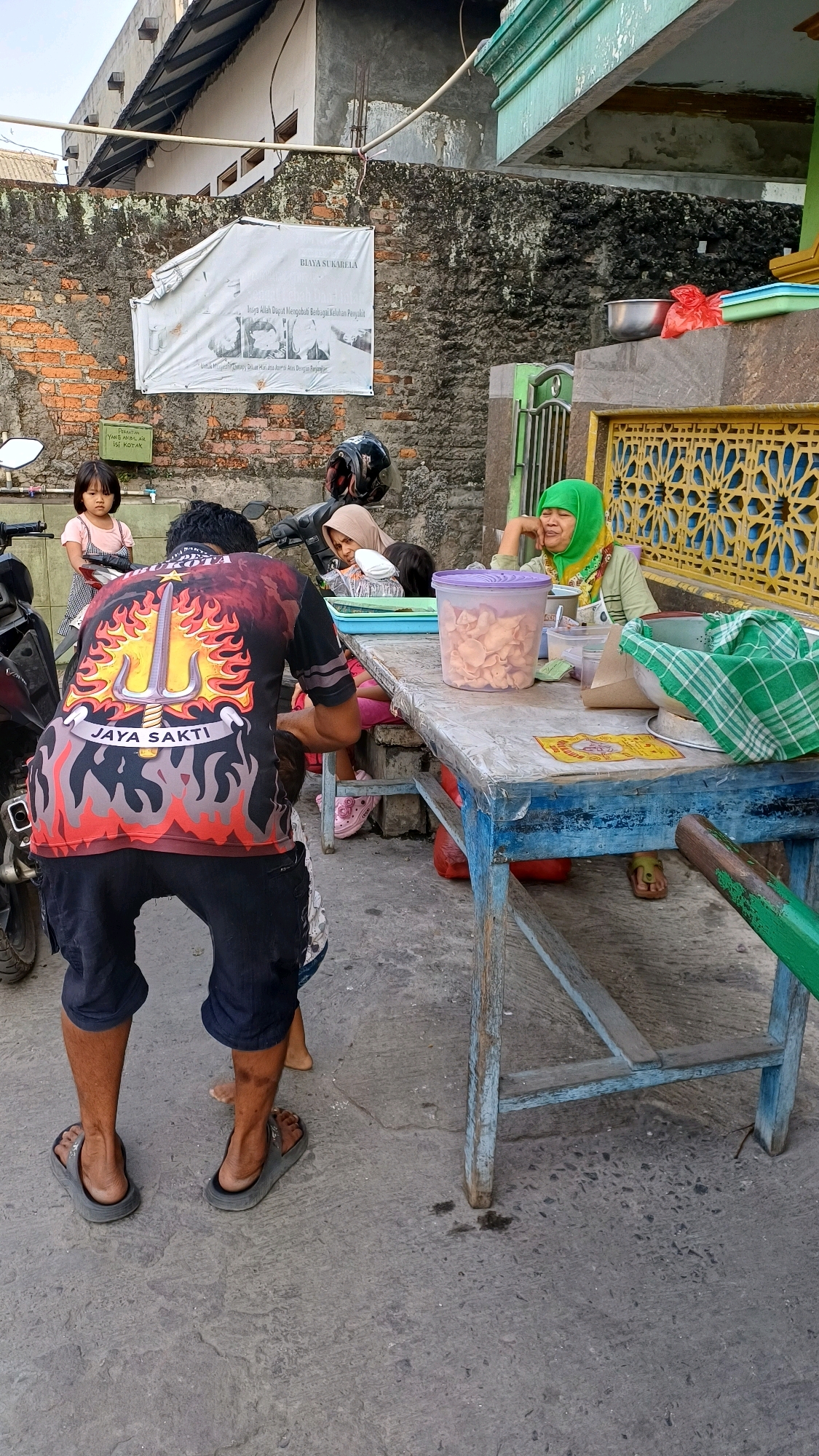 A scene unfolds on a concrete ground in front of a brick wall in Citeureup, Indonesia. In the foreground, a man wearing a black t-shirt with a flaming trident design and dark shorts bends down. He is wearing dark flip-flops. A young girl in a pink dress stands to the left, facing the camera. Behind her, a motorcycle is parked.
To the right, a long, weathered blue table is set up. On the table, a woman wearing a green hijab and a light green top sits, leaning her head on her hand. She appears to be looking to her left, with a slightly unhappy expression. Next to her, a clear plastic container is filled with light-colored chips. A green and white checkered cloth is draped over a bowl on the table.
The environment suggests an outdoor setting, possibly near a roadside stall or a community gathering area, given the presence of the table, food items, and the casual attire of the people. The brick wall behind the subjects displays a white banner with text, partially obscured. The building facade above the wall is visible, showing architectural details and exposed pipes.
