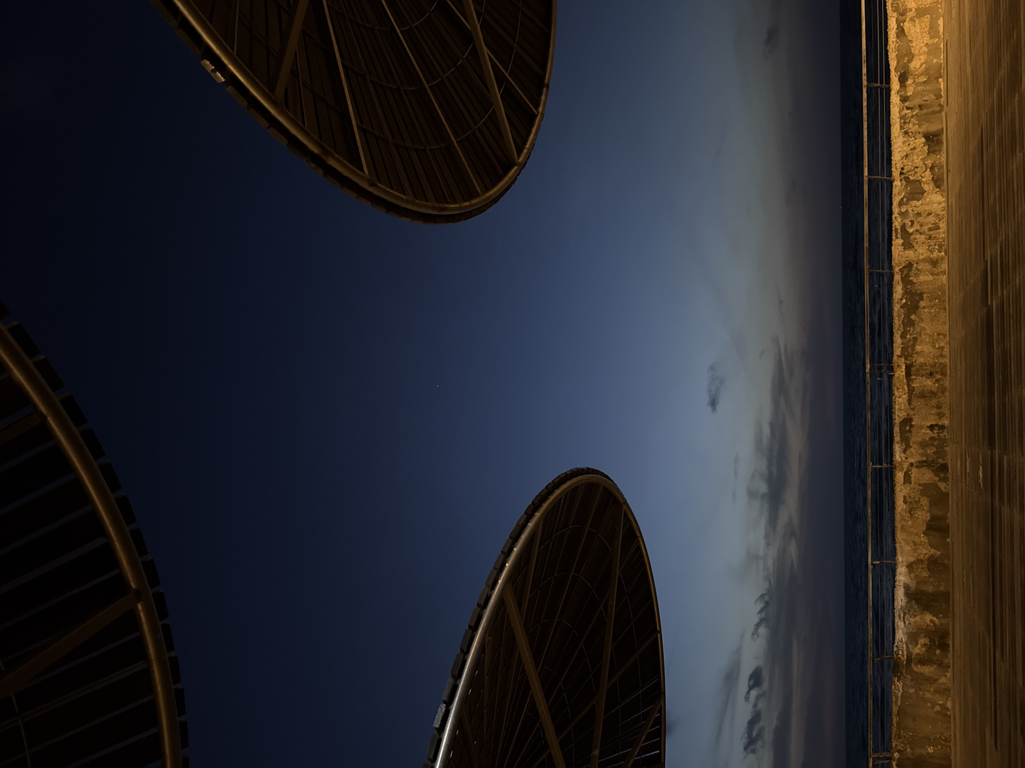 The image shows an abstract, low-angle shot, possibly taken at night or dusk. Two circular, caged structures with a metallic frame are visible on the top left and bottom left of the frame. The background appears to transition from a deep blue sky on the left to a lighter sky with clouds toward the right. On the right side of the image is a wall or edge, which includes stone and possibly a wooden walkway. The edge is uneven, and lit by a dim, warm-toned light. The image is rotated, and depicts elements of a cityscape or seaside location, potentially in Tel-Aviv, Israel. The presence of the seaside wall and the round structures suggest a seaside walkway or park with architectural lighting elements, with the sea and sky in the background.