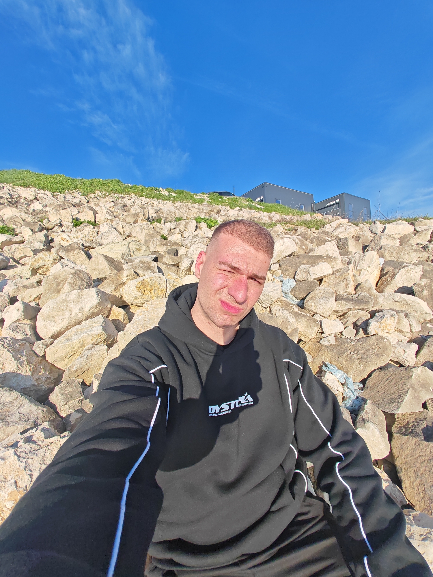 A man in a black hoodie is sitting on a rocky slope on a sunny day. The rocks are light-colored and vary in size. Green vegetation is visible on the slope above the rocks. In the background, there are two gray buildings with flat roofs. The sky is blue with wispy clouds. The man is looking at the camera with one eye squinted. The location is likely near the city of Shumen, Bulgaria, based on the buildings and landscape.