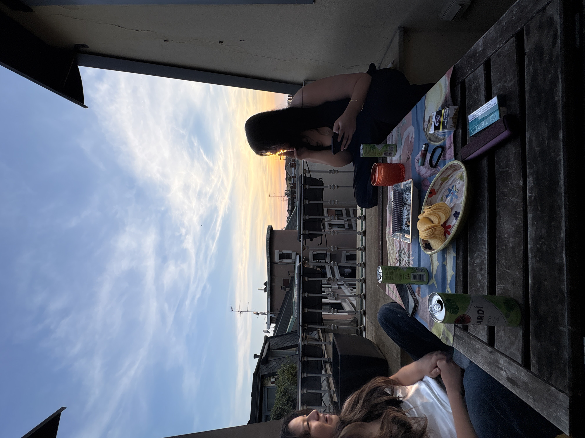 This image, rotated 90 degrees clockwise, captures a serene evening scene on a city balcony or rooftop terrace at sunset. Two women are relaxing, likely enjoying the view and company. One woman with long dark hair is seated on the floor, engrossed in her phone, her profile silhouetted against the bright horizon. Partially visible in the foreground is another woman with brown hair, also seated, looking away from the camera.
A wooden slatted table in front of them holds various items. A colorful mat covers a section of the table, displaying a bowl overflowing with potato chips, several green cans of beverages (at least two identified as Bacardí Mojito), an orange cup, a lighter, and a hair tie. A packet of tobacco products, labeled "TEREA" and featuring the Italian health warning "Questo prodotto da tabacco danneggia la tua salute" ("This tobacco product harms your health"), is also present.
The backdrop reveals the rooftops and buildings of a city, specifically Milan, Italy, under a beautiful sunset sky. The horizon glows with vibrant yellow and orange hues, blending upwards into a soft blue with scattered wispy clouds. The distinct European architecture of the buildings and a prominent television antenna on a distant roof confirm the urban setting. The overall atmosphere is one of relaxation and casual enjoyment during the golden hour.