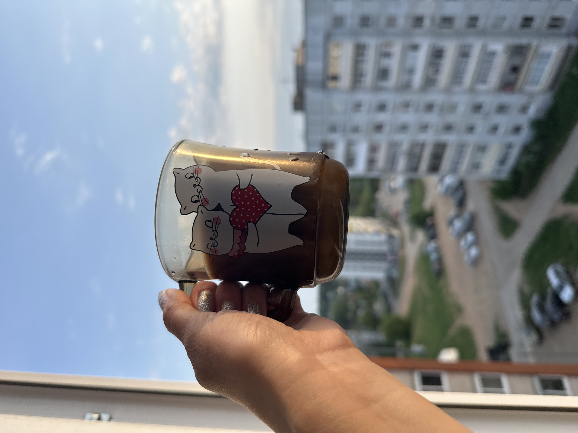 A hand holds a glass mug filled with a brown liquid, possibly coffee or tea. The mug has a cartoon drawing of two white cats with a red, heart-shaped patterned fabric. The background shows an urban scene with buildings, including a tall, multi-story apartment building with many windows. In the distance, there are parked cars and green areas, likely trees or grass. The sky is visible and partly cloudy. The photograph was likely taken in the city of Chaikovsky, Russia.