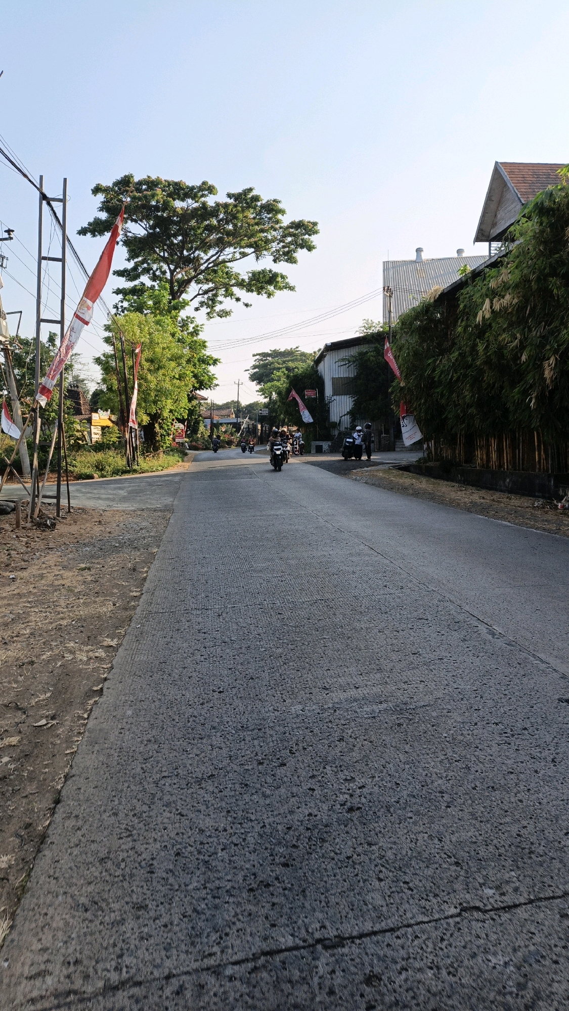 The image depicts a straight, paved road in Bandengan, Indonesia, during daylight. The road is grey asphalt with visible cracks and a textured surface. The left side of the road is bordered by a dirt and gravel shoulder with some dry grass. On the left, there is a metal pole with power lines attached, and several red and white Indonesian flags are attached to the pole and visible on flagpoles along the roadside, suggesting a national celebration or event. To the left and further down the road, there are various buildings and trees, some with green foliage.
On the right side of the road, there is a dense line of tall, green bamboo plants. Behind the bamboo, there are buildings with tiled roofs and metallic structures.
Several motorcycles are visible on the road, moving in both directions. In the distance, a group of motorcycles are traveling together. Two individuals on motorcycles are also visible closer to the right side of the road. The sky is clear and light blue, indicating fair weather. The overall scene suggests a rural or suburban setting with typical Indonesian road features and decorations.