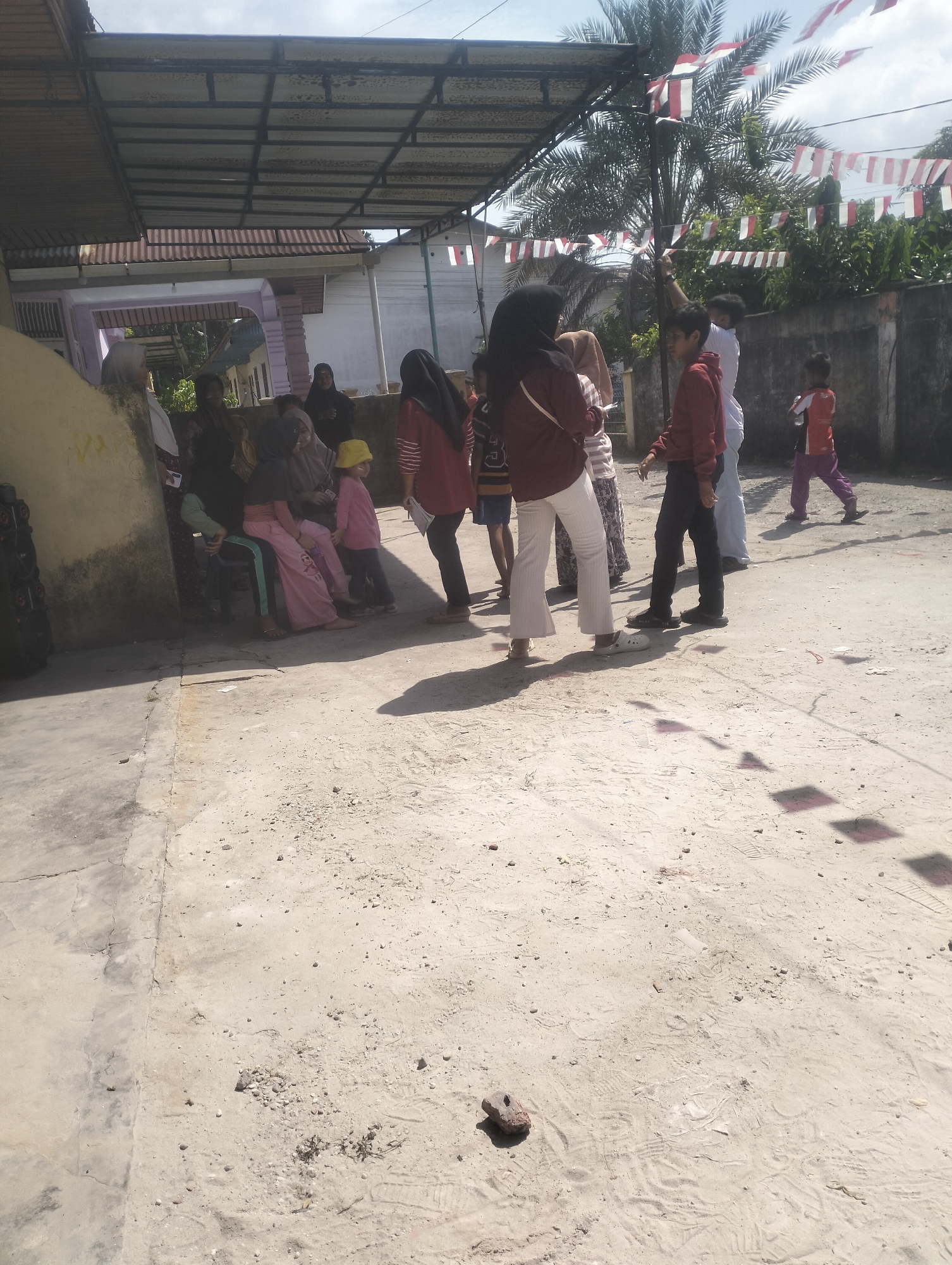 The image depicts an outdoor scene with a group of people gathered under a covered structure. In the background, palm trees and festive red and white Indonesian flags strung across the area suggest a celebration or national holiday. Several individuals are seated on chairs or on the ground, while others are standing. The ground is paved with concrete and appears to be a courtyard or open area.
The scene appears to be located in Dumai City, Indonesia, indicated by the presence of Indonesian flags and the style of the buildings and architecture. The overall atmosphere seems casual and communal, with people interacting and observing the surroundings.