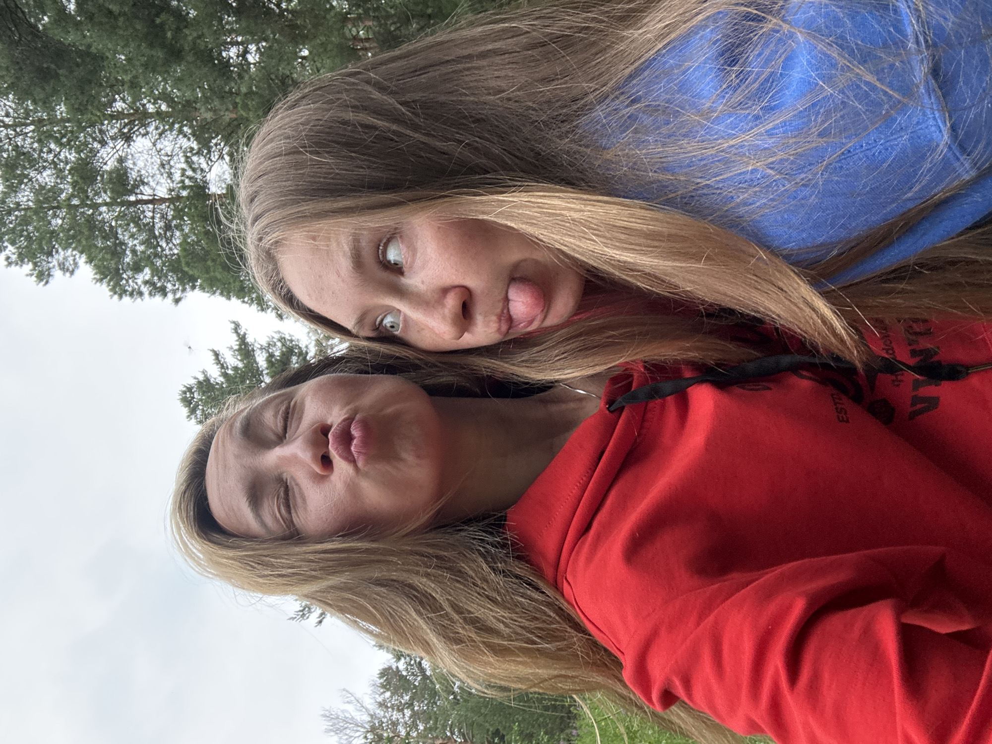 The media file is a selfie taken outdoors with two young women. The image is oriented sideways. The woman in the foreground is wearing a red hooded sweatshirt. Her eyes are closed, and her lips are puckered as if for a kiss. Her hair is long and light brown. Behind her, another young woman is visible, wearing a blue hooded sweatshirt. She has light brown hair and is making a silly face with her tongue sticking out to the side and her eyes wide open. The background consists of green trees and a cloudy sky, suggesting an overcast day. The overall atmosphere appears casual and playful, as indicated by the facial expressions. There is some faint text visible on the red sweatshirt, but it is largely illegible. The location is identified as Прикамский, Russia.