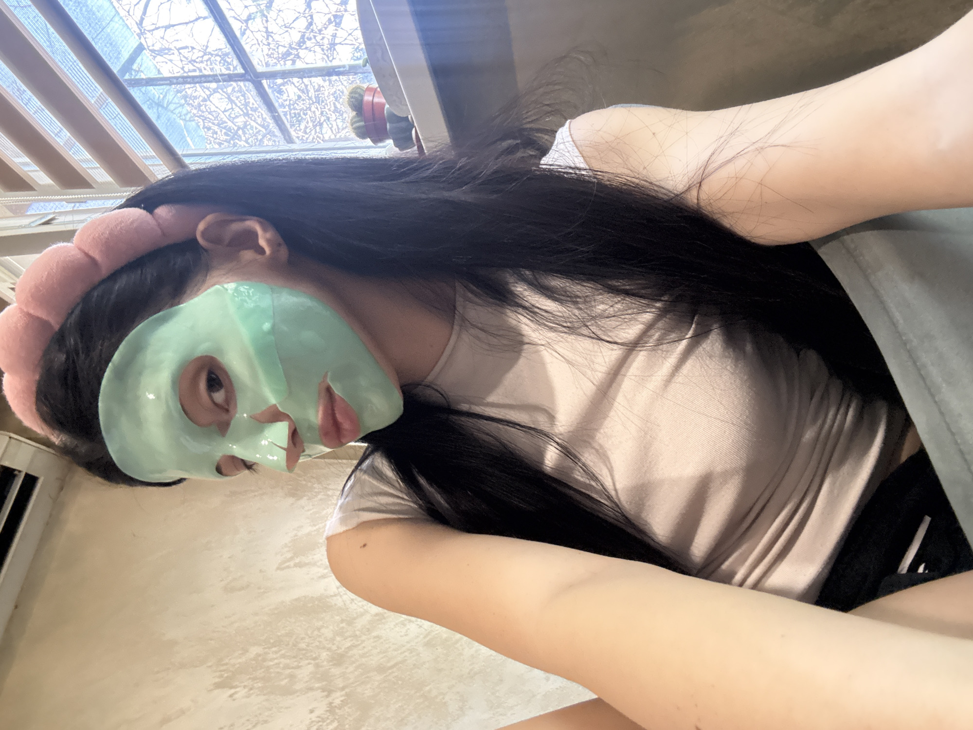 A person with a green sheet face mask and a pink headband is visible in the image. The person has long, dark hair and is wearing a light pink t-shirt. Black clothing is also visible. The background shows a window with blinds, and an interior wall. The image is likely taken indoors. The location in Sofia, Bulgaria cannot be determined from the image.