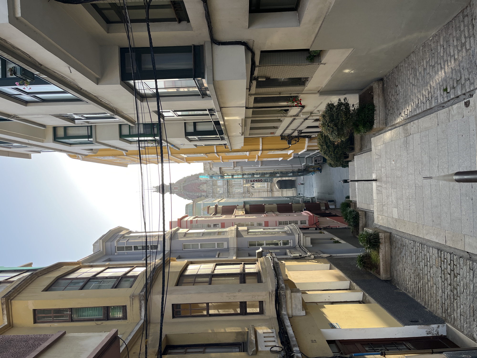 The image captures a narrow street in A Coruña, Spain, lined with buildings of varying heights and colors. The buildings are a mix of yellow, white, pastel blue, and pink, with several windows and balconies visible. Power lines crisscross overhead, adding to the urban feel. The street itself is paved with light-colored rectangular stones, with a darker cobbled stone pathway along the sides. In the distance, the ornate dome of a building is visible. There is sparse vegetation in the form of small bushes planted along the street. There is a sign for "PRADO" hanging above the street. A couple of people can be seen walking in the distance. The sun appears to be shining brightly, as evidenced by the glare in the sky.
