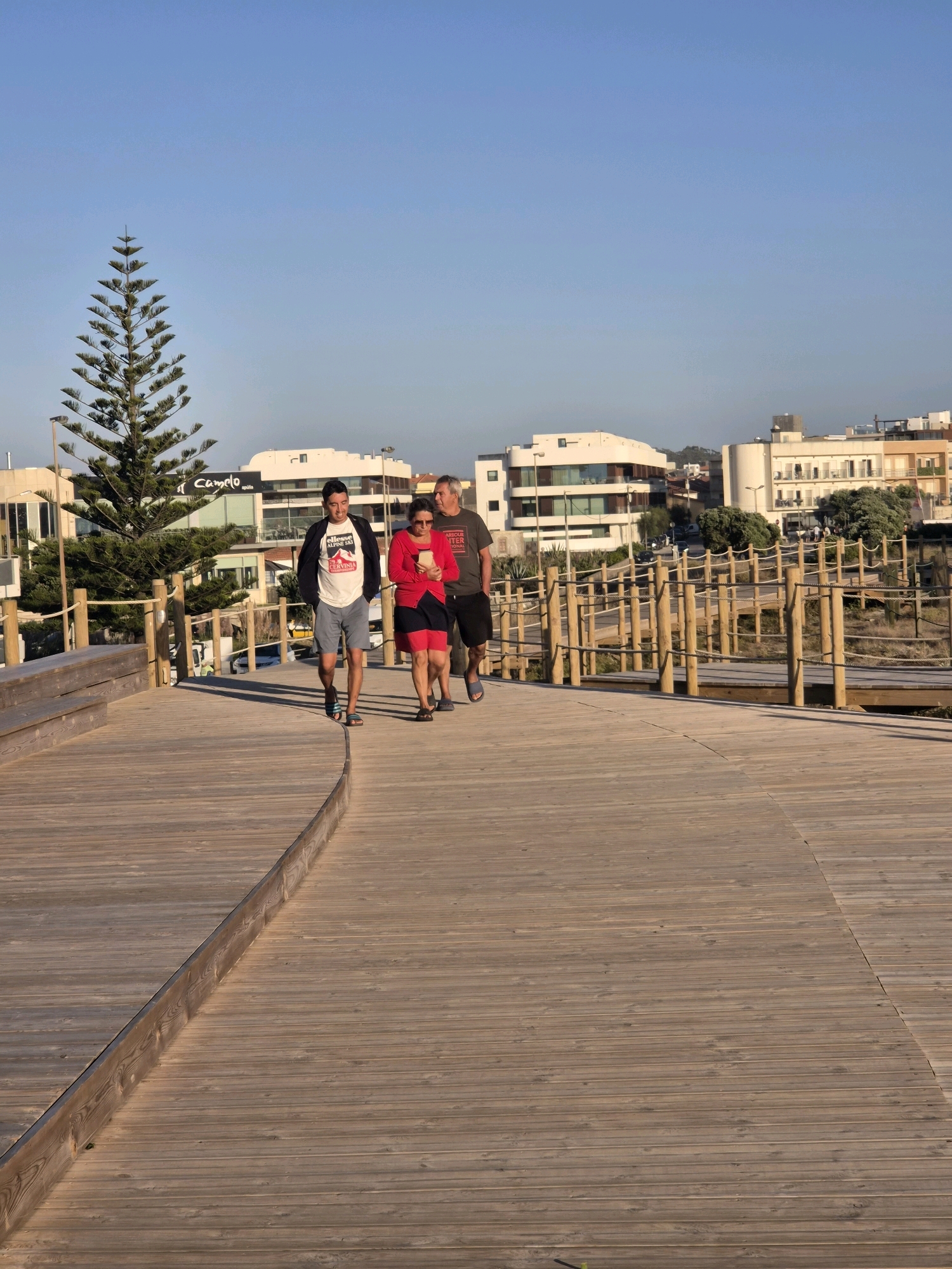 A low-angle shot captures three people walking on a wooden boardwalk in Esposende, Portugal. The sun is low in the sky, casting long shadows and creating a warm, golden light. The sky is clear and blue, suggesting fair weather.
In the foreground, the wide wooden boardwalk curves to the right, its planks showing a warm, sun-bleached tone. A darker wooden border lines the inner edge of the curve. To the left, a wooden bench is visible. A tall, slender Norfolk pine tree stands on the left side of the boardwalk, its branches reaching upwards.
The three individuals are walking away from the camera, towards the background. The person in the middle, a woman, is wearing a red top and a dark skirt with a red stripe. She is looking at her phone. To her left, a man in a white t-shirt with a graphic print, dark shorts, and a black jacket walks ahead. To the woman's right, another man in a dark t-shirt and dark shorts follows. All three appear to be casually dressed.
In the background, several modern buildings with white facades and rounded architectural elements are visible, suggesting a developed coastal area. A sign with the word "Camelot" is visible on one of the buildings. Wooden fencing with rope details lines the sides of the boardwalk, separating it from the sandy or scrubby terrain beyond. The overall scene evokes a relaxed atmosphere, likely a pleasant walk along the coast during the late afternoon or early evening.