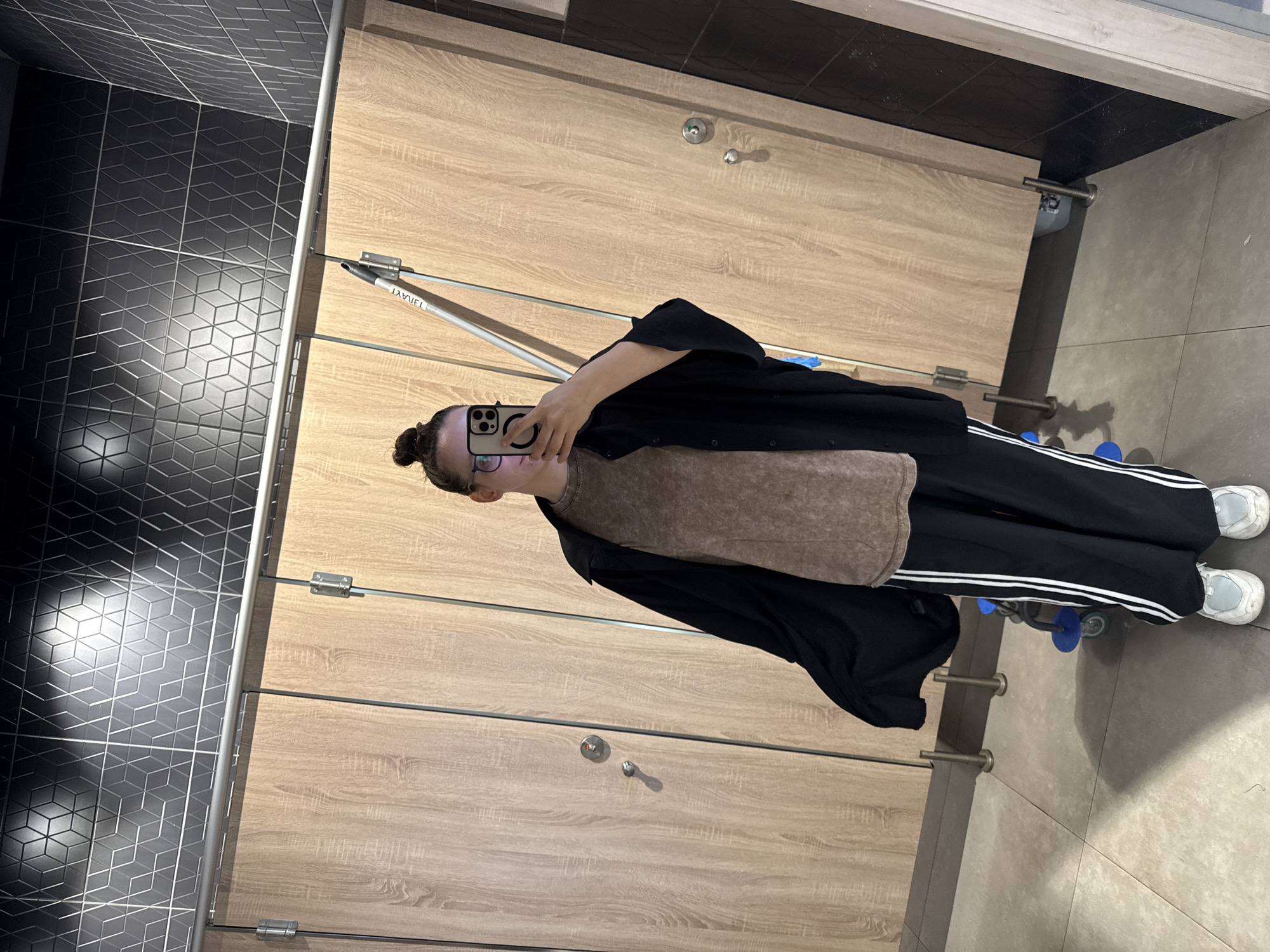 A person is standing in a restroom in Gil'deevo, Russia, taking a selfie in front of a row of wooden stall doors. The person has dark hair pulled back in a bun and is wearing glasses, a brown shirt, a black cardigan, and black sweatpants with white stripes. They are holding a cell phone up to their face to take the picture. The restroom has dark gray tile on the wall and light gray tile on the floor. The stall doors are light wood with metal hardware.