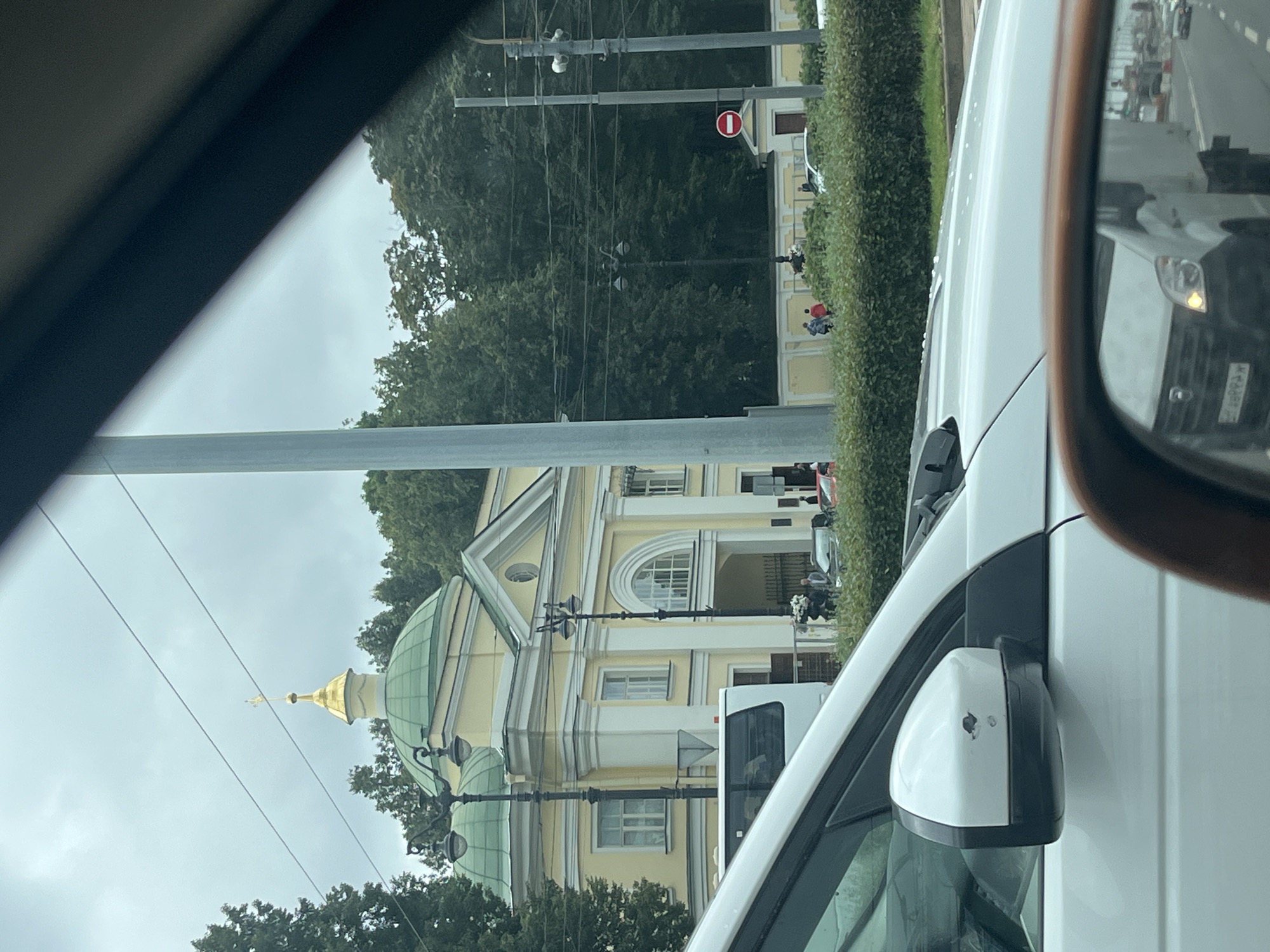 The image captures a view through a car window in Saint Petersburg, Russia. In the background, a light yellow building with green domes and a golden cross on one of the domes is visible, likely a church. The building features classical architectural elements, including columns and arched windows. Trees surround the building, and a hedge runs along the front. Several people are seen walking on the sidewalk in front of the building. A white bus is parked near the building. The foreground is dominated by the white car from which the photo is taken, with its side mirror and part of the hood visible. The sky is overcast. The scene appears to be on a city street with traffic and utility poles.