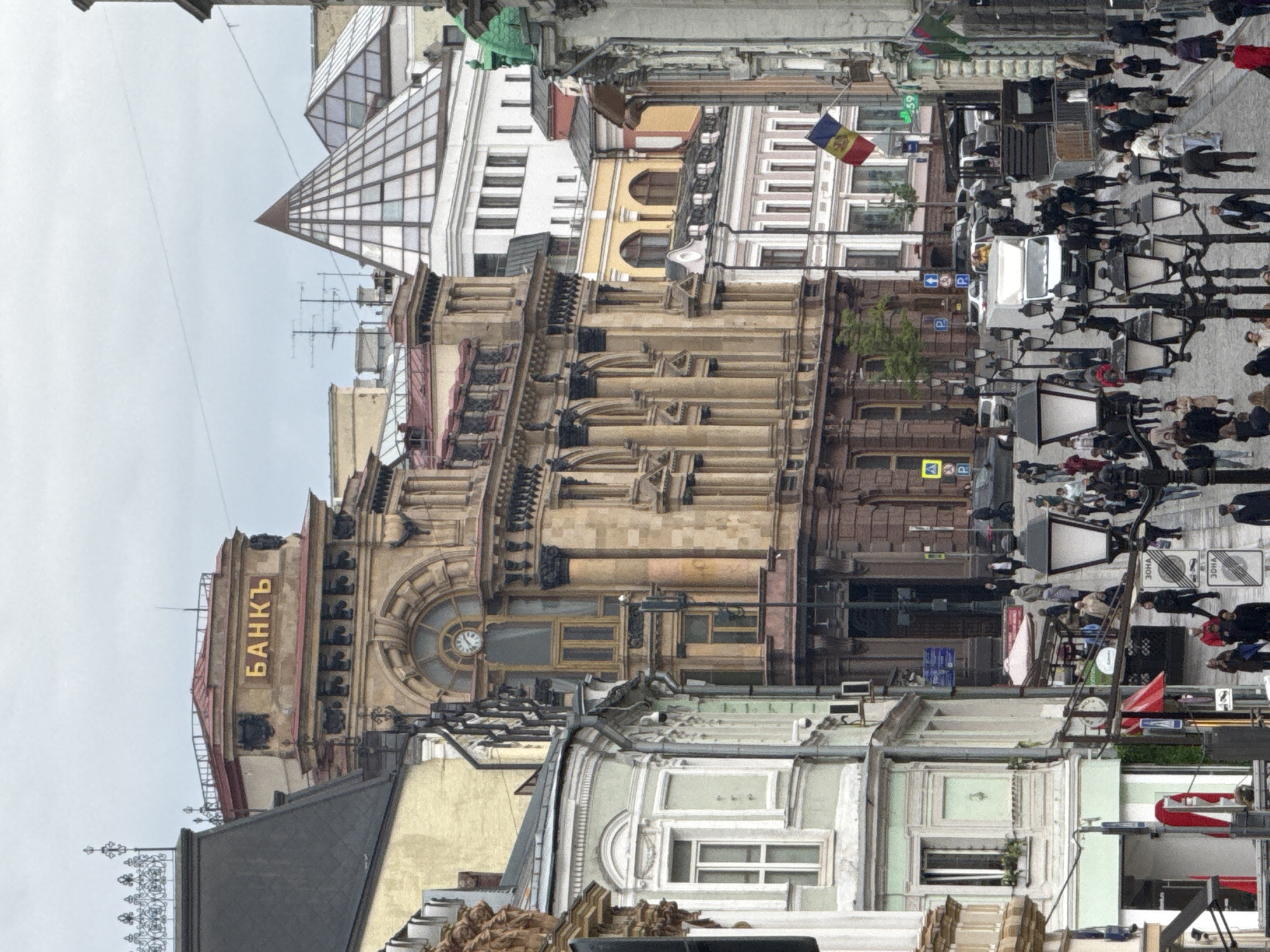 The image captures a vibrant street scene in what is identified as Moscow, Russia, characterized by a mix of historic and contemporary architecture under an overcast sky.
In the foreground and central midground, a grand, multi-story building dominates, featuring intricate classical architectural details including numerous fluted columns, ornate window frames, and decorative cornices. Prominently displayed in gold Cyrillic letters on its facade is the word "БАНКЪ" (Bankъ), an older spelling for "Bank." A large, circular clock is visible on an upper level of this building. To the left, a more modern structure with a distinctive pyramidal glass roof rises into the cloudy sky. Various other buildings, ranging from ornate older styles to more contemporary designs, line the street.
The street level is bustling with numerous people walking and congregating, suggesting a busy urban environment. Many individuals are dressed in dark or neutral-colored clothing, indicative of daytime activities. Rows of black street lamps with multiple light fixtures illuminate the pedestrian area. A line of electric scooters or bikes is visible, likely available for public use, reflecting modern urban transportation trends. Several cars are also present on the street, along with various street signs, including blue parking signs with a white 'P' and signs with Cyrillic text, one of which clearly reads "ЗОНА" (Zone). A small flag, featuring red, yellow, and blue vertical stripes with an emblem in the center (identifiable as the flag of Moldova), hangs from a building in the midground. The overall impression is that of a lively urban commercial or historical district during the daytime, though the lack of harsh shadows suggests an overcast day.