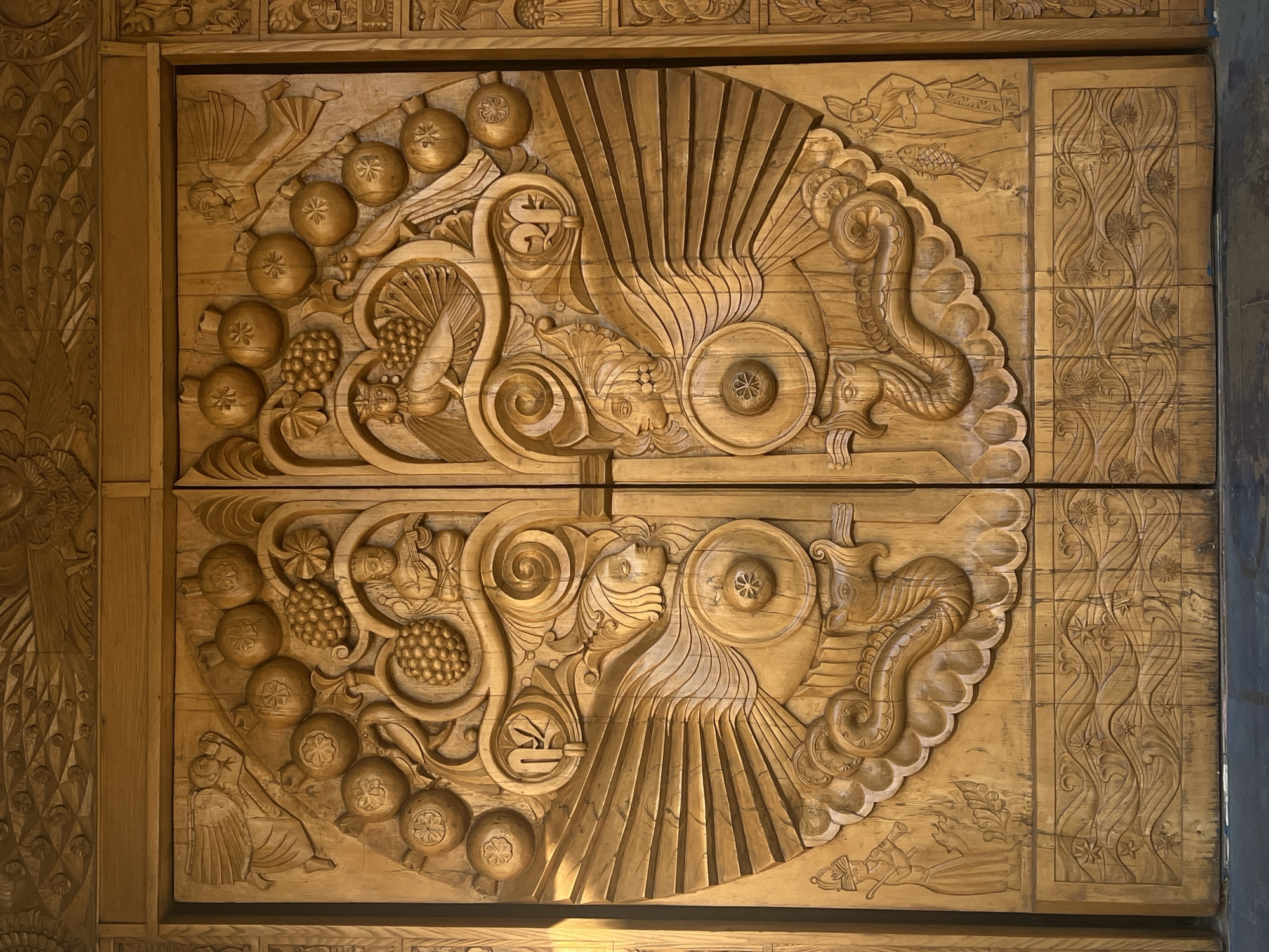 This image depicts a close-up of an ornate, carved wooden door or panel. The carving features a symmetrical design divided across the two halves of the door, with elements including pomegranates, grapes, scrolls, fan-like patterns, and stylized animal figures resembling dragons or seahorses. The wood is light in color, and the carving is detailed with sharp lines and subtle shading. The exact location and city are unknown.