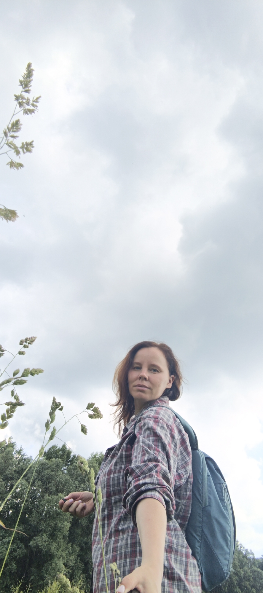 A woman with brown hair and a plaid shirt stands outdoors, with a backpack on. She is looking at the camera. The background is a cloudy sky with some green trees and grass. The location is Garevaya, Russia.