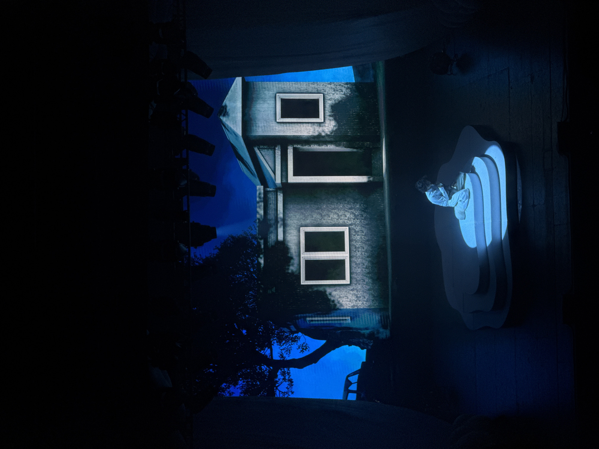 A dimly lit theatrical scene displays a woman posed on a stepped, cloud-shaped platform illuminated by a blue spotlight. To the left, a backdrop suggests a cityscape at night, featuring the silhouette of a tree branch against a building with illuminated window frames. Stage lighting equipment is subtly visible at the edges of the scene. The overall atmosphere is artistic and theatrical. There is no evidence of Los Angeles.