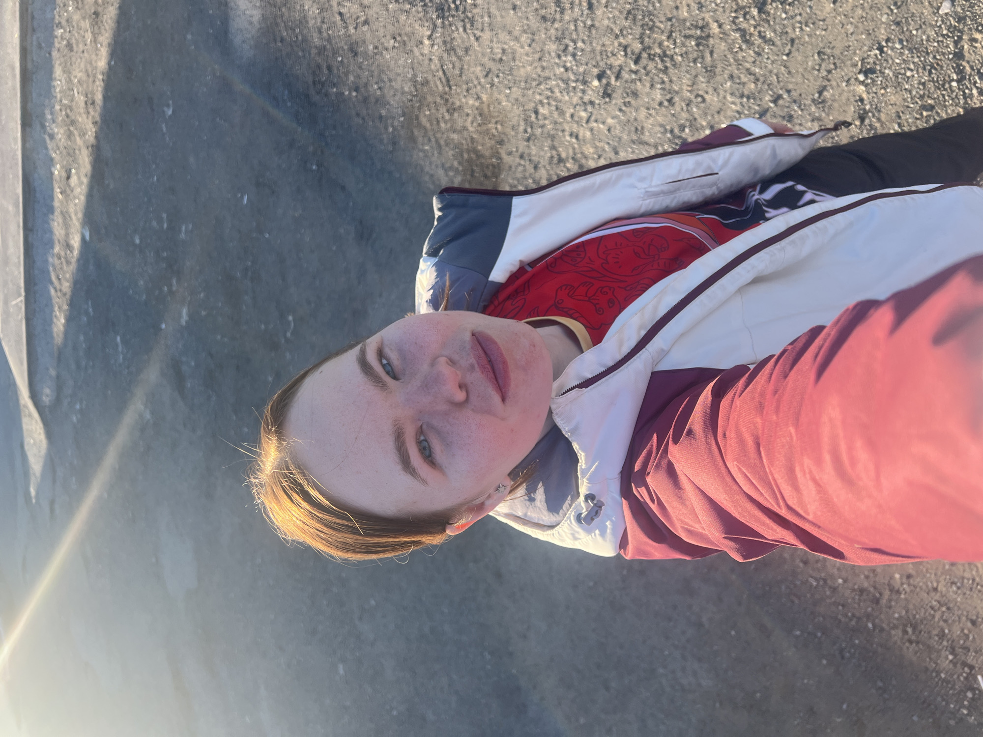 A person is standing outdoors on a paved surface, likely asphalt or concrete, in direct sunlight. The individual is wearing a red patterned shirt beneath a partially unzipped jacket with white, gray, and maroon panels. The person is fair-skinned with light hair that has some sunlit highlights. Their face is tilted upward towards the camera. Shadows cast by nearby structures are visible on the left side of the image. There is no discernible information in the image to determine the location in Nizhnie Yaki, Russia.