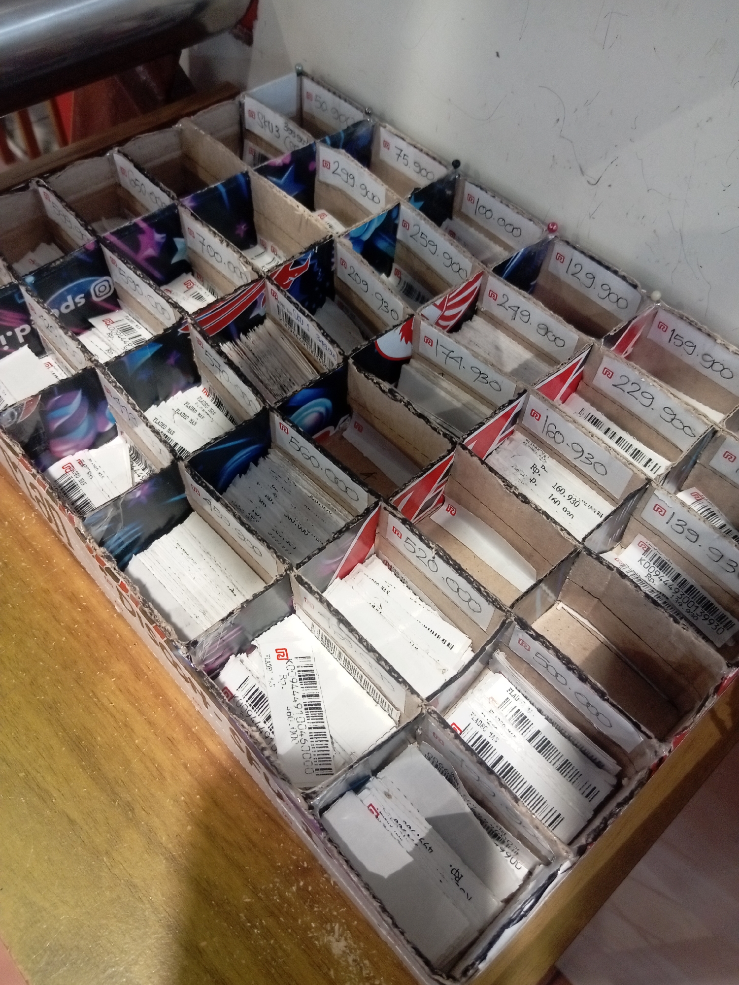 The image shows a close-up, high-angle shot of a compartmentalized cardboard box filled with stacked, small, white rectangular items. Many of these items appear to be vouchers or gift cards, with some displaying colorful graphics and branding such as "Instagram" and a stylized "S". Several of the items have handwritten or printed numerical values, often preceded by "Rp.", suggesting they are in Indonesian Rupiah. Examples of these values include "75.000", "100.000", "259.900", "129.900", "174.930", "229.900", "159.900", "139.930", "560.000", and "520.000". The box itself is made of corrugated cardboard and has dividers creating a grid of approximately 5x7 compartments. The lighting appears to be natural or from an overhead source, casting soft shadows. The background is out of focus and shows what appears to be a light-colored, textured wall and a dark, metallic surface above the box. The overall scene suggests an organized collection of financial instruments or promotional items. Based on the presence of "Rp." and the visible branding, the location is likely Batam, Indonesia.