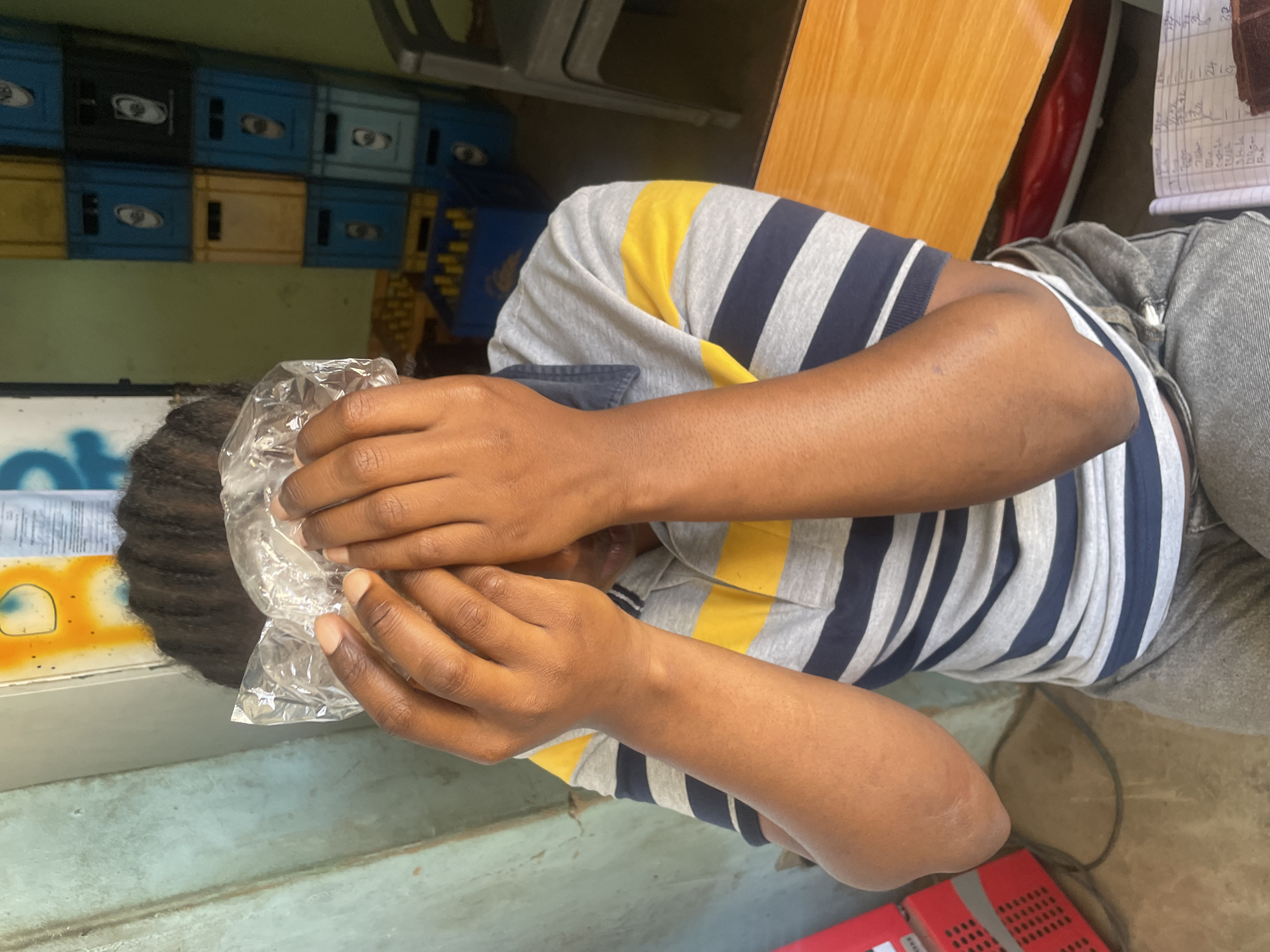 A person sits in what appears to be a small store or shop. They are holding a plastic bag to their face, obscuring it from view. They wear a striped t-shirt with grey, navy blue, and yellow stripes, as well as light-colored denim pants. In the background, there are stacks of plastic crates in blue and yellow. The store seems to be in Jos, Nigeria based on the local product in the background.