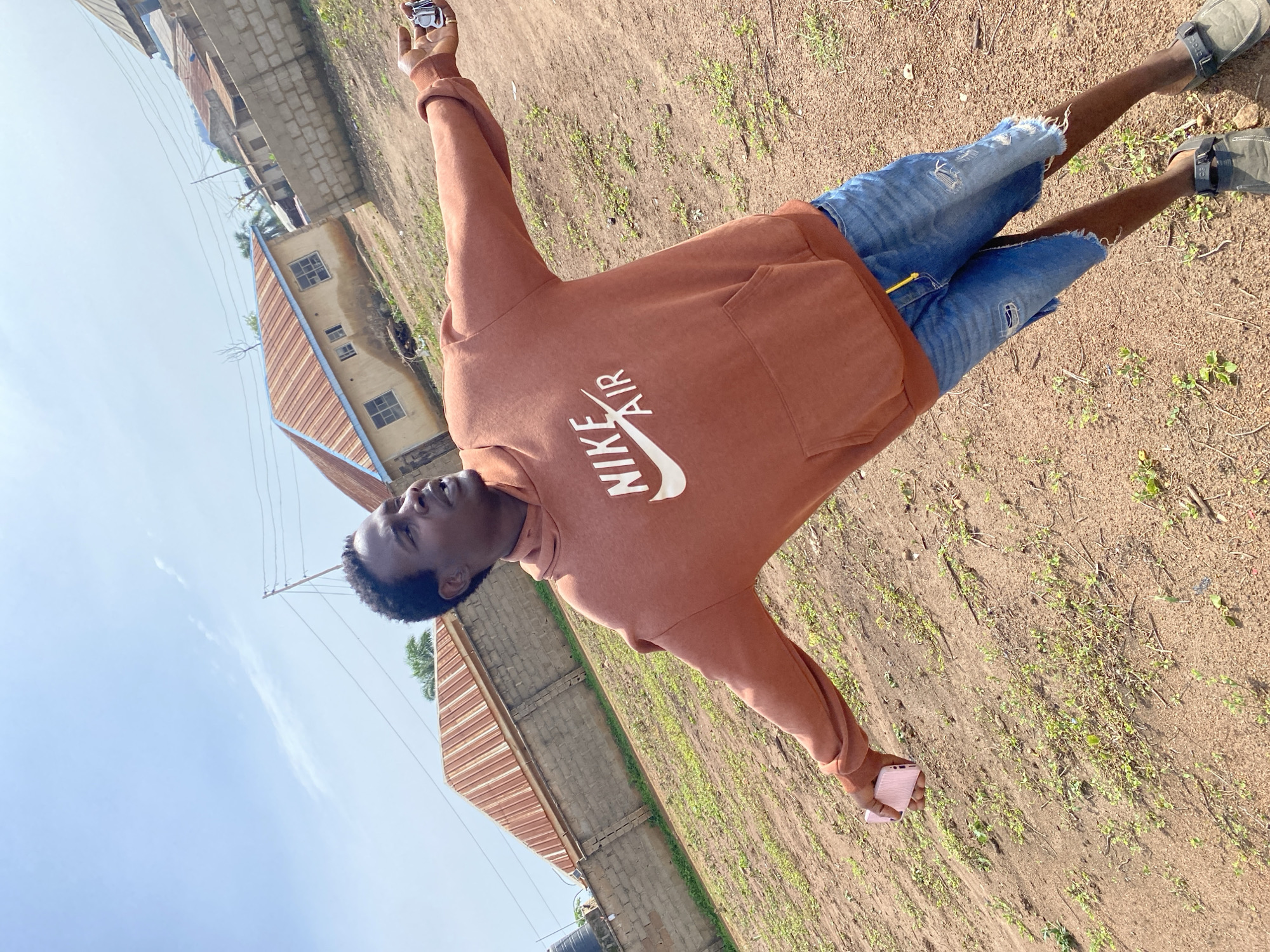 A young man, with dark skin and short, black hair, stands outdoors in what appears to be a residential area. He is wearing a rust-colored hoodie with "NIKE AIR" printed in white on the chest, and distressed denim shorts. His left arm is extended outwards, with his hand holding a pink smartphone. His right arm is also extended, and his hand is open. The background consists of simple, low-rise buildings with corrugated metal roofs, a brick wall, and a dirt ground sparsely covered with green vegetation. The sky above is a clear blue with some wispy clouds. The overall scene suggests a casual, outdoor setting.