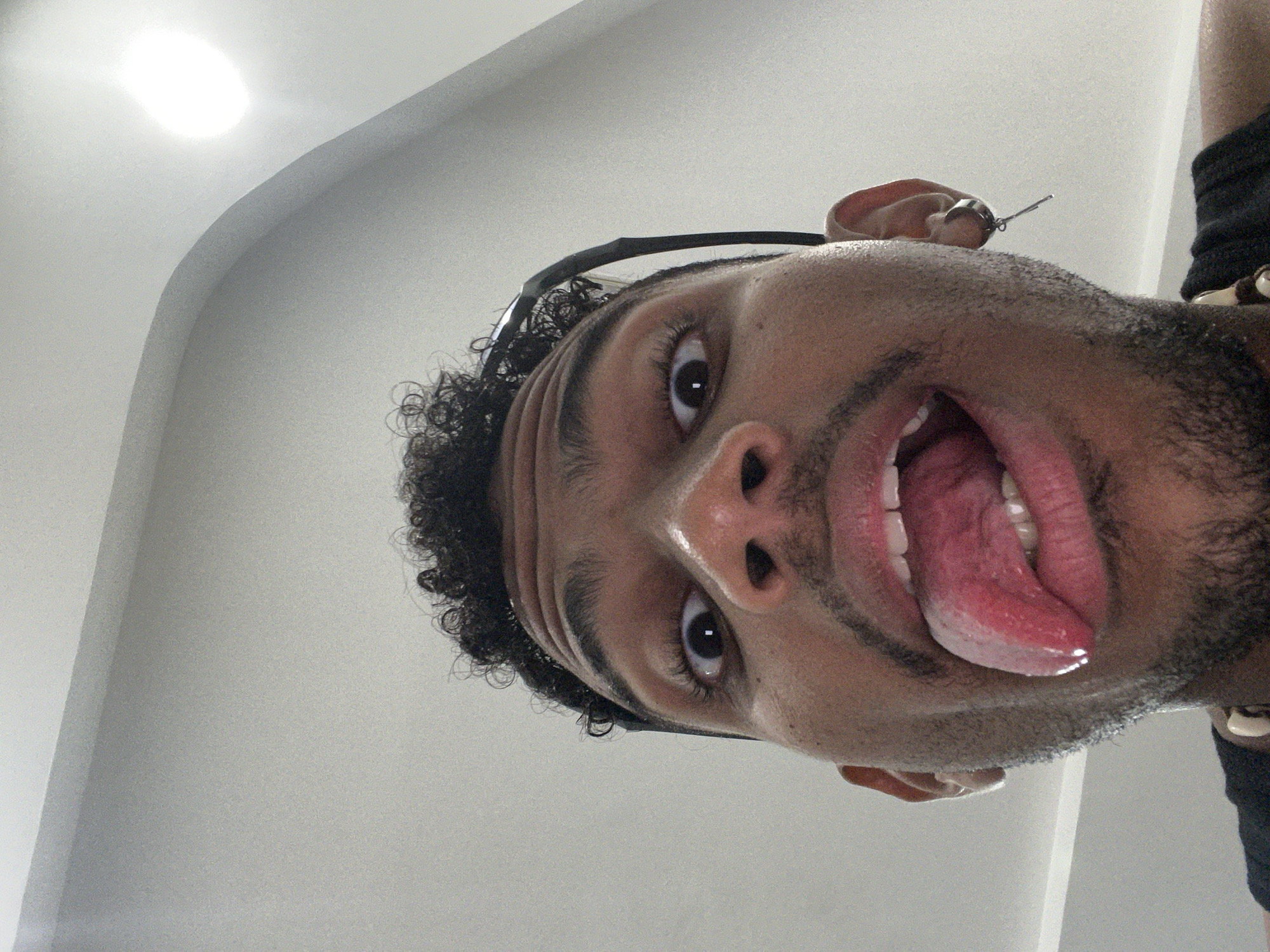 A low-angle close-up shot of a dark-skinned male with curly black hair, stubble, and a partially visible beard. He is wearing glasses on top of his head and a black shirt with a beaded accessory. His mouth is open with his tongue extended. The background is a plain white wall with a light source. The location of this image is unknown and there is no explicit evidence to suggest this photo was taken in Porlamar, Venezuela.