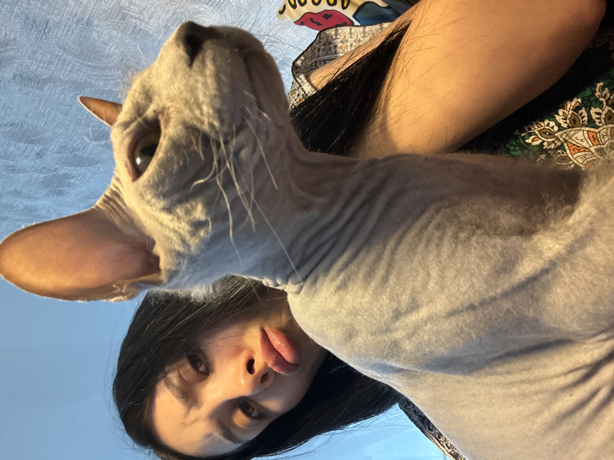 A woman with long dark hair is lying on her back, her face upturned toward the camera with her tongue sticking out. Next to her, a hairless cat is positioned similarly, looking upwards. The woman's arm is visible, resting on a patterned textile that appears to be part of a bedspread or clothing. The background is somewhat blurred, showing a textured wall or surface. Location is unknown and cannot be derived from the image. The image appears to be taken indoors.