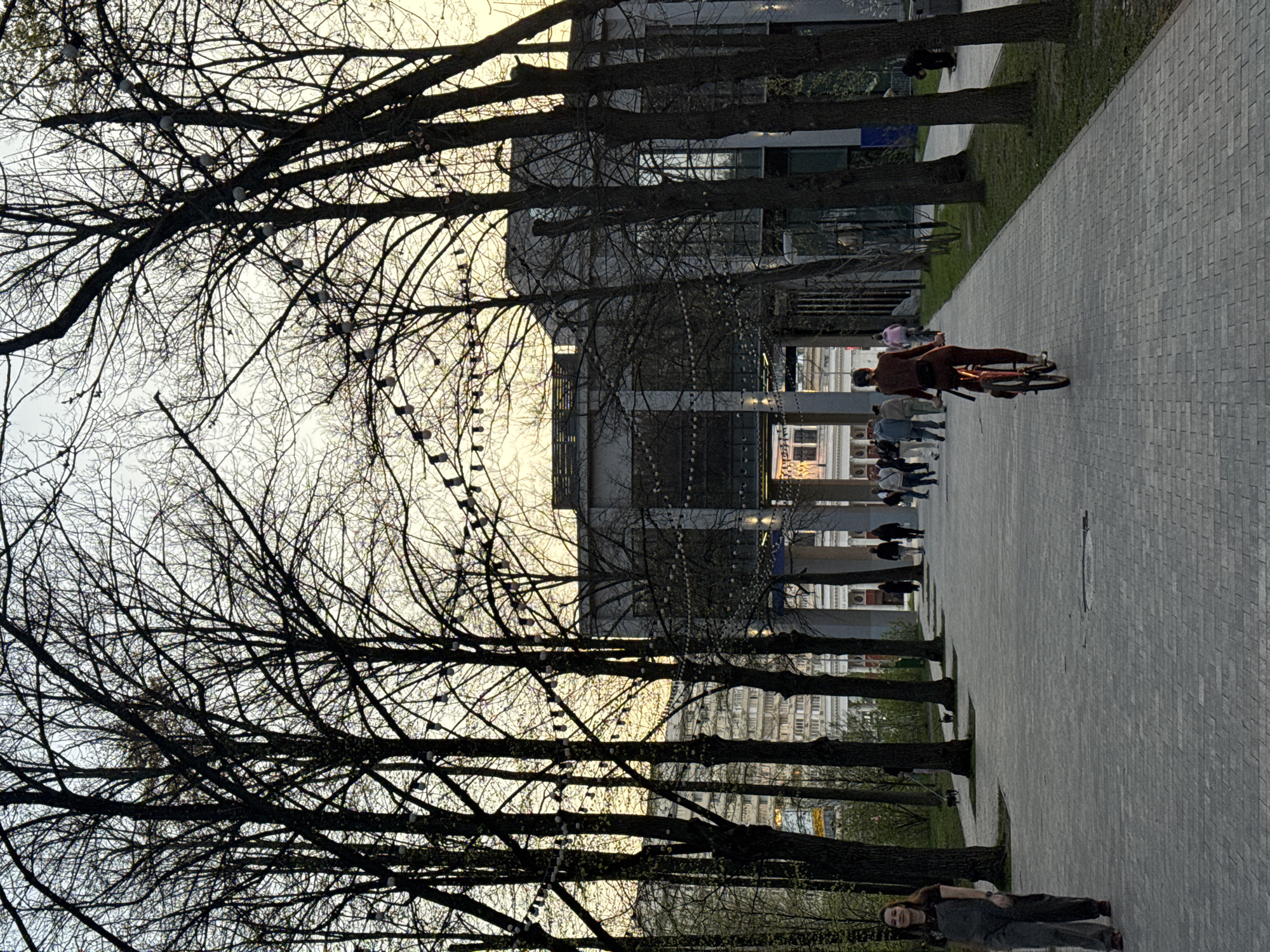 The image captures a scene along a pedestrian walkway in Moscow, Russia. The walkway is paved with gray rectangular stones and lined on one side by a row of tall, bare trees. Strings of lights are suspended between the trees. Behind the trees and lights, a modern building is visible, constructed of glass and metal, with other buildings in the background. Several people can be seen walking and riding bicycles along the walkway. In the foreground, a woman lies on her side on the paving stones. The sky is overcast, suggesting either early morning or late afternoon.