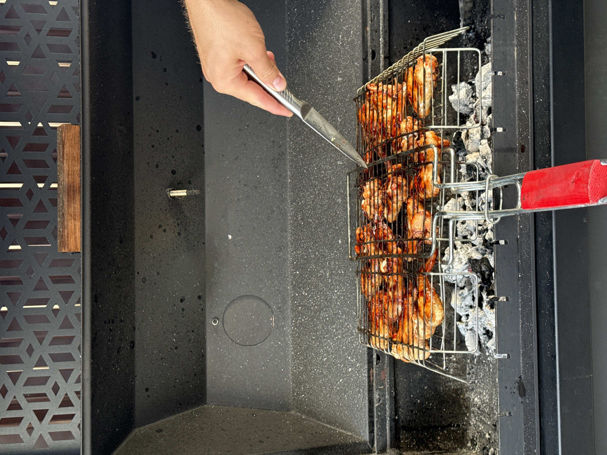 A close-up, eye-level shot shows a person's right hand holding a silver-handled knife, with the blade pointed towards a grill basket filled with glistening, glazed chicken pieces over hot coals. The grill basket is positioned inside a dark, black metal barbecue. The barbecue has a decorative metal grate with a geometric pattern on the top back wall, and a wooden block rests on it. The environment appears to be outdoors, given the natural light and the context of a barbecue. There is no discernible information about the specific location within Starokorsunskaya, Russia, or any other identifying features that would narrow down the precise setting beyond it being a barbecue area. The focus is on the act of preparing food, with the chicken appearing to be marinated or basted with a dark sauce. The coals beneath the grill are visible, covered in white ash, indicating they are hot and actively producing heat. The overall scene suggests a culinary activity, likely a meal preparation.