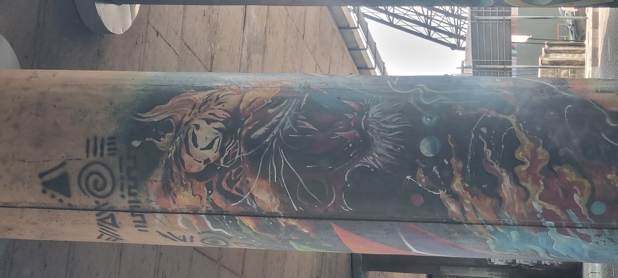 A horizontal concrete beam, possibly part of an underpass or elevated structure, is adorned with vibrant graffiti art. The artwork spans a significant portion of the beam, depicting an energetic and abstract scene against a dark, almost cosmic background.
On the far left of the mural, black stencil-like patterns, including a triangle with dots and a prominent spiral, are visible. Moving right, the painting transitions into a striking, fierce-looking animal face, possibly a tiger or lion, rendered in shades of orange, brown, and black stripes, with its mouth open. This animal imagery blends into abstract fiery or energetic formations.
Further along, the mural features a spiky, organic shape with radiating white tendrils, which might suggest a sun, star, or an explosive event. Beside it, several spherical objects resembling planets or moons in various sizes and shades of blue, white, and red are scattered across the dark expanse, accompanied by white splatters and streaks, evoking a cosmic atmosphere. The rightmost section of the painting continues with dynamic, flame-like or liquid patterns in warm hues of red, orange, and yellow, with touches of blue and white, suggesting elemental forces or abstract energy.
Above the painted beam, a bright, overcast sky is visible, partially obscured by dark metal lattice work, indicative of an overhead structure such as a bridge or roof support. Faint outlines of other distant structures can also be seen. The concrete beam itself shows signs of wear, with visible textures and minor imperfections, providing a gritty contrast to the detailed artwork. The overall impression is a blend of tribal motifs, powerful animal symbolism, and cosmic or elemental abstraction. No legible text is visible within the artwork, only abstract symbols.