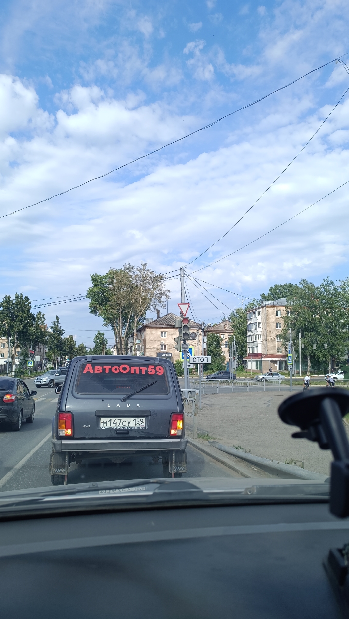 A dark gray LADA Niva is stopped at a traffic light, viewed from behind. On its rear window, red lettering reads "АвтоОпт59". The car's license plate is M147CY 159. In front of the LADA, other vehicles are stopped. Beyond the intersection, there are multi-story buildings, trees, and a cloudy blue sky. A traffic light is red, and a stop sign is visible. The scene appears to be in an urban environment in Chaikovsky, Russia.