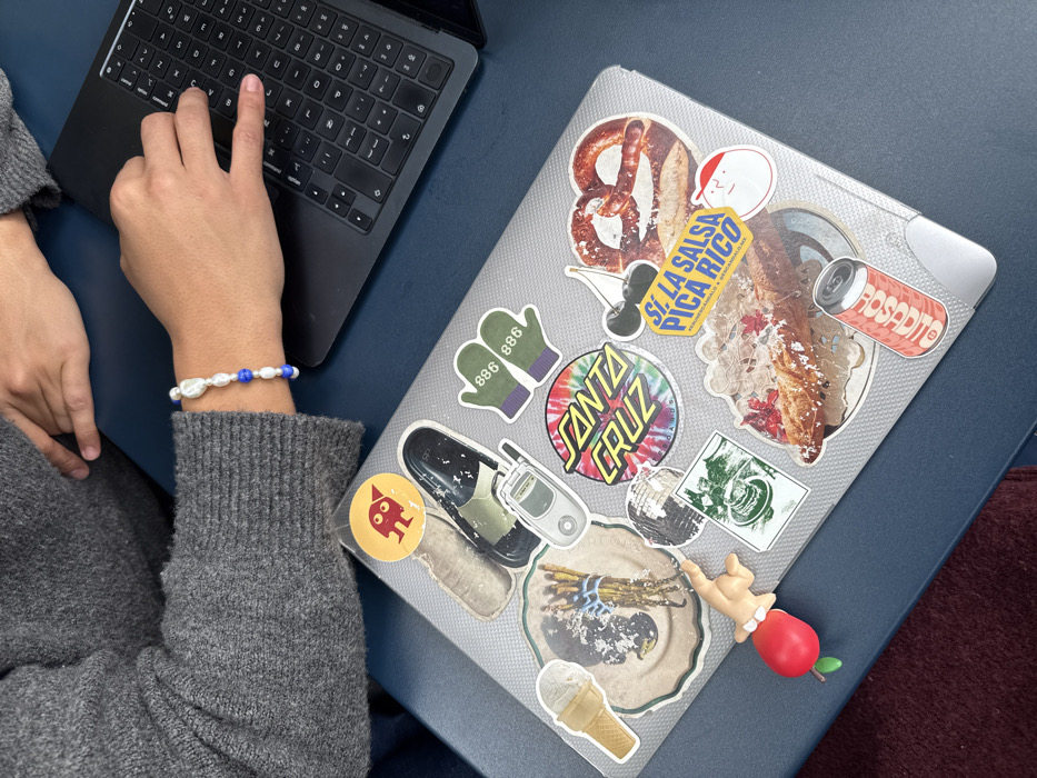 A person types on a black laptop, wearing a gray sweater and a bracelet of white and blue beads. Next to the laptop is a silver laptop adorned with numerous stickers: a pretzel, mittens labeled "886 886," a Santa Cruz logo, a vintage phone, an ice cream cone, a "Sí La Salsa Pica Rico" sticker, a Rosadito soda can, and a figure resembling a baby with a red apple body.