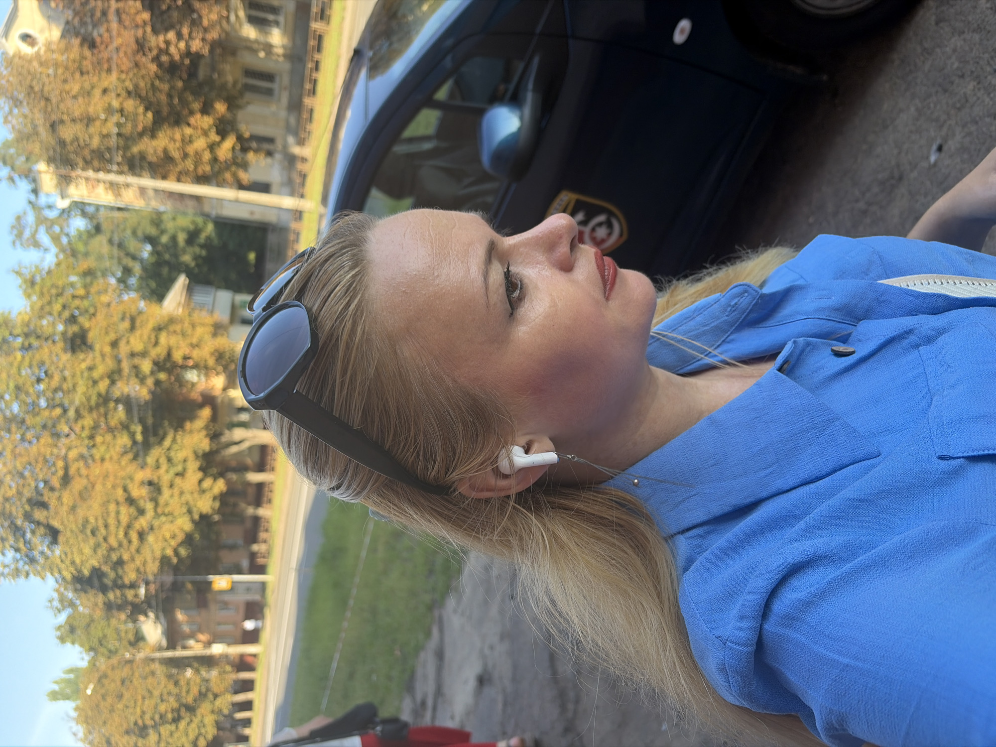 An outdoor medium close-up shot shows a fair-skinned blonde woman with blonde hair and sunglasses pushed back on her head. She is wearing a bright blue collared shirt with buttons. The woman has on a white wireless earbud in her right ear, with a silver thread hanging from it. She is looking to the right of the frame. Behind her, a dark blue vehicle with a circular emblem on the door is visible. Trees with yellowing leaves and buildings line a street in the background.