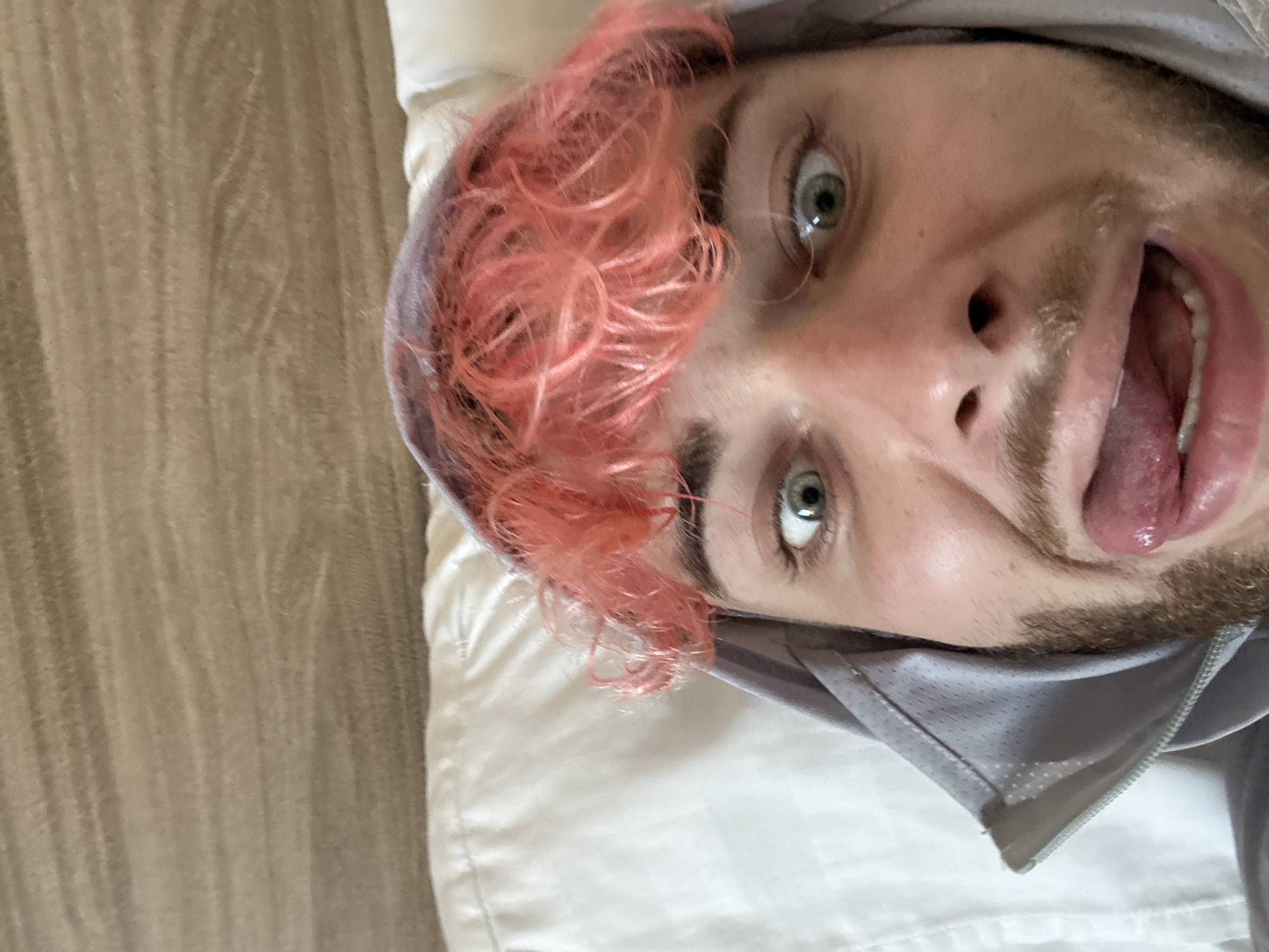 A close-up, rotated view of a person lying down. The person has fair skin, a beard, and a small amount of facial hair. The person's hair is styled into tight curls and is dyed a light pink color. The person's eyes are open and appear light in color. Their tongue is extended out of their mouth. They are wearing a grey hooded sweatshirt, and they are lying on a white pillow. The background consists of light brown flooring. There is no indication in the image that this is in Đà Nẵng, Vietnam.