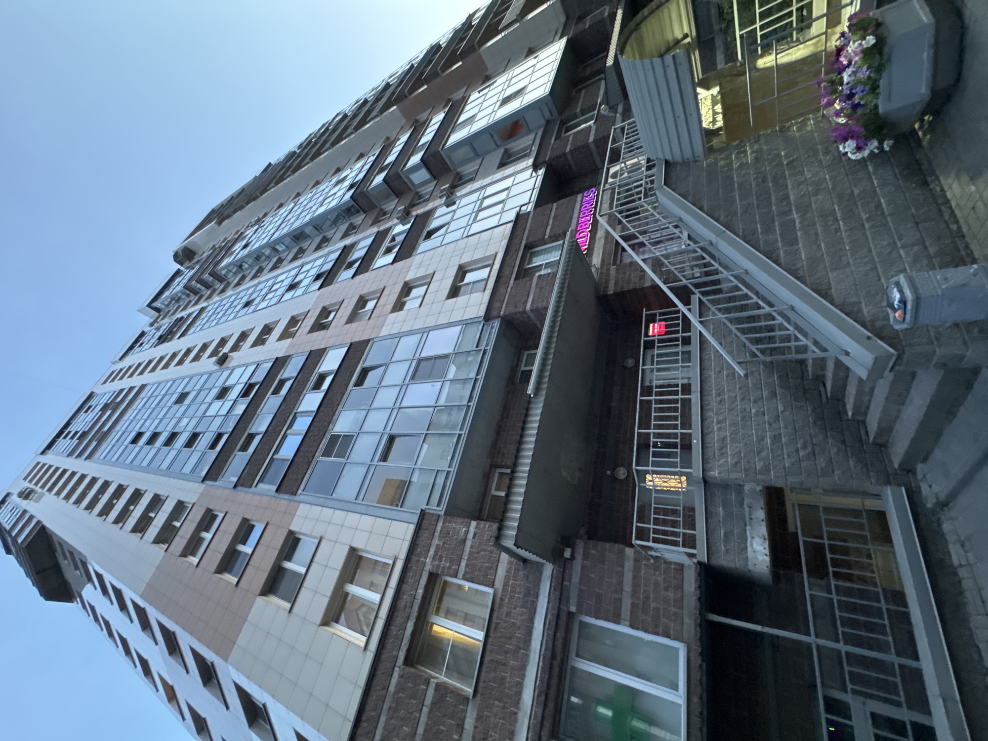 An exterior, low-angle, wide shot captures the facade of a multi-story apartment building under a clear blue sky in an urban setting. The building is tall and slender, with a mix of light beige and brown brickwork, punctuated by rows of windows and glass balconies. The lower section of the building features a covered entrance area with a staircase leading up to a railing. A sign with pink neon lettering, partially visible, reads "ildberries". To the right of the staircase, a large concrete planter overflows with a colorful arrangement of purple, white, and pink flowers. A gray trash receptacle stands to the left of the planter. The image appears to be taken during the late afternoon or early evening, as suggested by the angle of the sunlight.