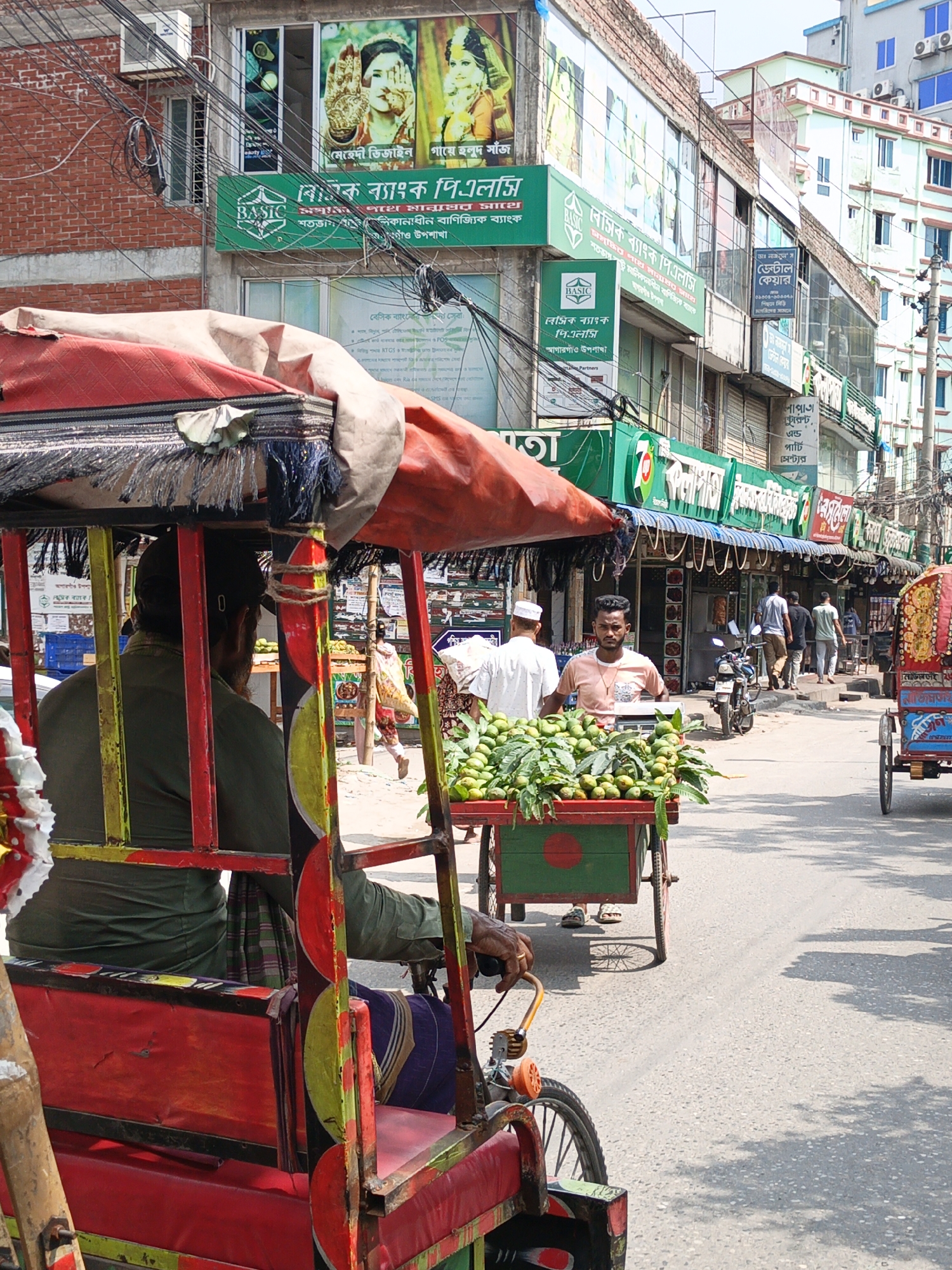 The image shows a street scene in an urban area. A brightly painted, red, yellow, and green rickshaw is in the foreground, with the driver wearing a green shirt and a lungi. Behind the rickshaw is a cart filled with green fruits, pushed by a man in a white skullcap and another man in a pink t-shirt. Pedestrians walk on the sidewalk in the background. Various businesses line the street, including one with a green sign advertising "BASIC Bank PLC" and another indicating a dental care service. Electrical wires are visible overhead. Buildings of varying heights and architectural styles make up the backdrop.