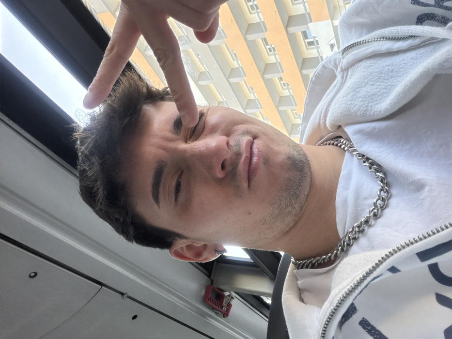 A young adult male, approximately 20-30 years old, with fair skin, short dark hair, and light facial stubble, is depicted in a close-up, selfie-style photograph. He is wearing a white t-shirt, a prominent silver-colored Cuban link chain necklace, and a white zippered hoodie. A small silver stud earring is visible in his left ear. His left eye is closed, and his right eye is partially open, with his lips slightly pursed in what appears to be a wincing or playful expression. His right hand is raised, with the index finger extended and touching the outer corner of his left eye, while other fingers form a V-shape.
The subject is seated inside a public transport vehicle, likely a bus or tram. The interior features a light-colored ceiling with dark circular elements and a matching side panel with a horizontal ridge. A red emergency or control device is partially visible mounted on the interior wall to the upper left of the subject.
Through the vehicle's window, which occupies the right side and upper portion of the frame, a multi-story building is visible under bright daylight. The building exhibits a repetitive architectural pattern with an orange-tan facade, featuring numerous white-framed rectangular windows and balconies across multiple floors. This type of residential or commercial architecture is characteristic of urban environments in Sofia, Bulgaria, establishing the location within the city of Sofia.