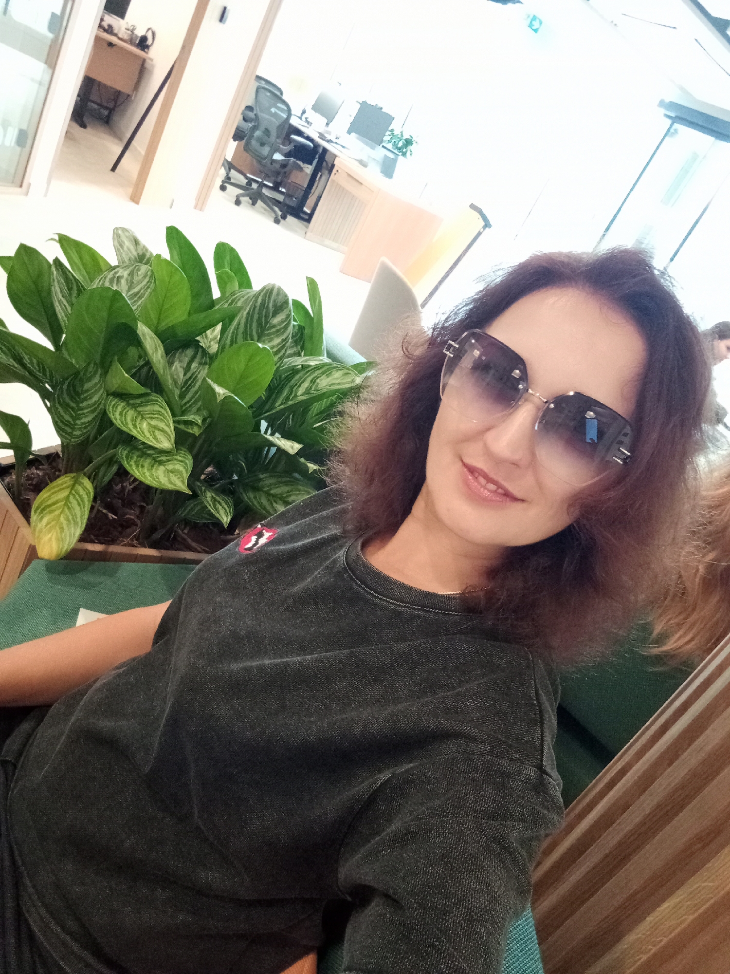 In an indoor office environment, a woman with dark, wavy hair and wearing large, fashionable sunglasses poses for a selfie. She is dressed in a dark, charcoal-colored t-shirt with a red and white emblem on the left breast. She is seated in what appears to be a teal-colored chair, next to a large potted plant with variegated green leaves. The background suggests an open-plan office with natural light streaming in. Visible are glass partitions, office furniture, and desks with computers, indicating a modern workspace. The scene does not provide any information to suggest it is located in the city of Riga, Latvia.