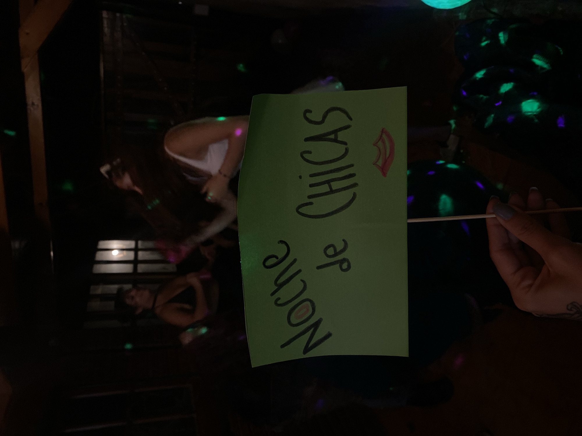 The image shows a dimly lit indoor scene, likely at a party or gathering. Two women are visible in the background. A green sign is held up, displaying the words "Noche de Chicas" (Girls' Night) in black marker, with a lipstick drawing near the word "Chicas." A hand holding the sign is visible on the right side of the frame. The scene is illuminated by green and blue light effects. The location is possibly in Monte Grande, Argentina.