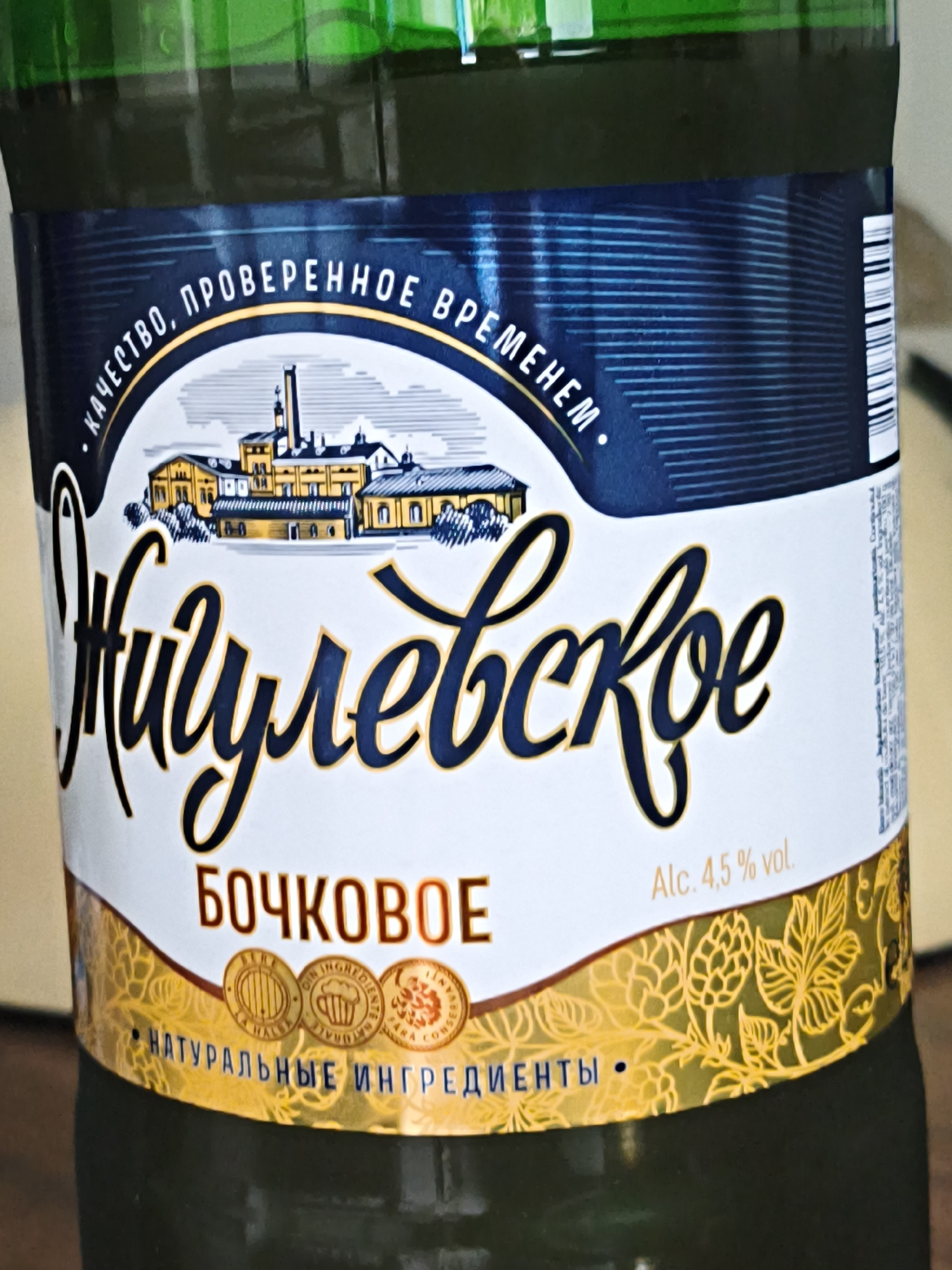 A close-up shot shows a portion of a dark green bottle with a label. The label has a white background with blue and gold accents. At the top of the label, there is text in a yellow arc that reads ". КАЧЕСТВО, ПРОВЕРЕННОЕ ВРЕМЕНЕМ.". Below this, there is an illustration of an old factory building. Dominating the center of the label is the word "ЖИГУЛЕВСКОЕ" in large, stylized blue script with gold outlining. Below this, in all caps and gold lettering, is the word "БОЧКОВОЕ". To the right of this, in smaller white text, it reads "Alc. 4.5% vol.". At the bottom of the label, a blue dotted line runs horizontally, and to the left of it, in blue text, it says "• НАТУРАЛЬНЫЕ ИНГРЕДИЕНТЫ •". The bottom portion of the label features a decorative gold pattern of hop leaves and cones. The image is taken from a slightly elevated angle, focusing on the label itself. The location in the city of Chișinău, Moldova cannot be determined from the provided image.