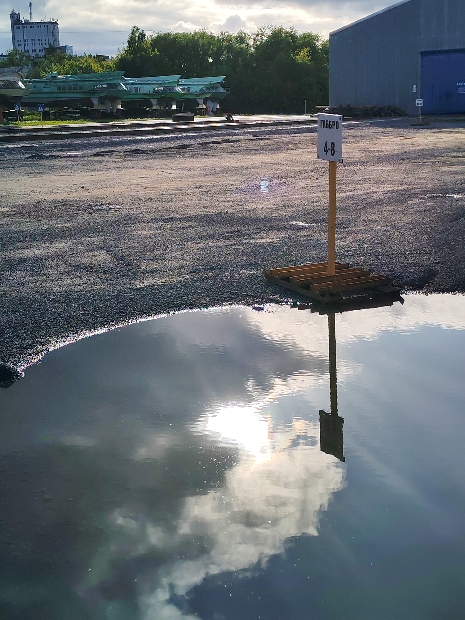 The image captures a sunlit industrial or storage area, seemingly after a recent rain, as evidenced by a large puddle in the foreground. The puddle reflects the sky, including clouds and the sun. A signpost with Cyrillic script "ГАББРО" and numbers "4-8" stands slightly off-center to the right, its base resting on a makeshift wooden platform. The signpost is reflected in the puddle. The ground is covered in gravel. In the background, there are several green structures, possibly vehicles or machinery, raised on supports. Further back, trees and a tall, blocky building are visible, suggesting an urban setting. To the right, a large, silver-colored building with a closed blue door is present. The lighting is bright, with harsh shadows indicating direct sunlight. The location is in the city of Unknown, Unknown.