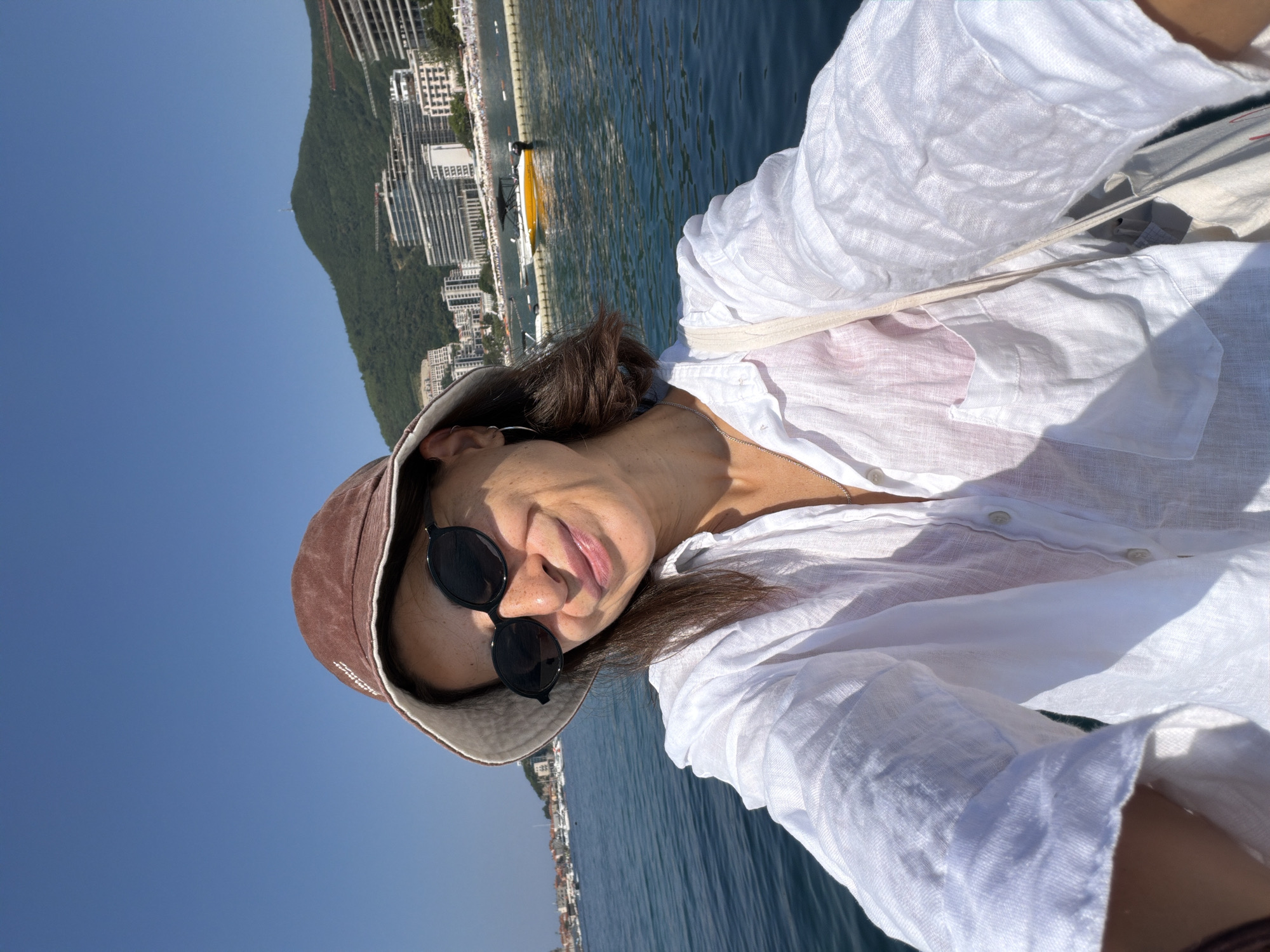 A woman is taking a selfie on a boat, with a city and a lush green mountain in the background. The sun is shining brightly, casting a clear shadow on her white linen shirt. She is wearing a brown bucket hat and dark sunglasses, which are perched on her nose. Her hair is dark brown and pulled back. The woman has a slight smile on her face, and her lips are slightly parted. The water is a deep blue, and the sky is a bright, clear blue, suggesting it is a sunny day. The city in the background appears to be built along the coastline, with numerous buildings visible. The overall impression is one of a relaxed and enjoyable outdoor experience.