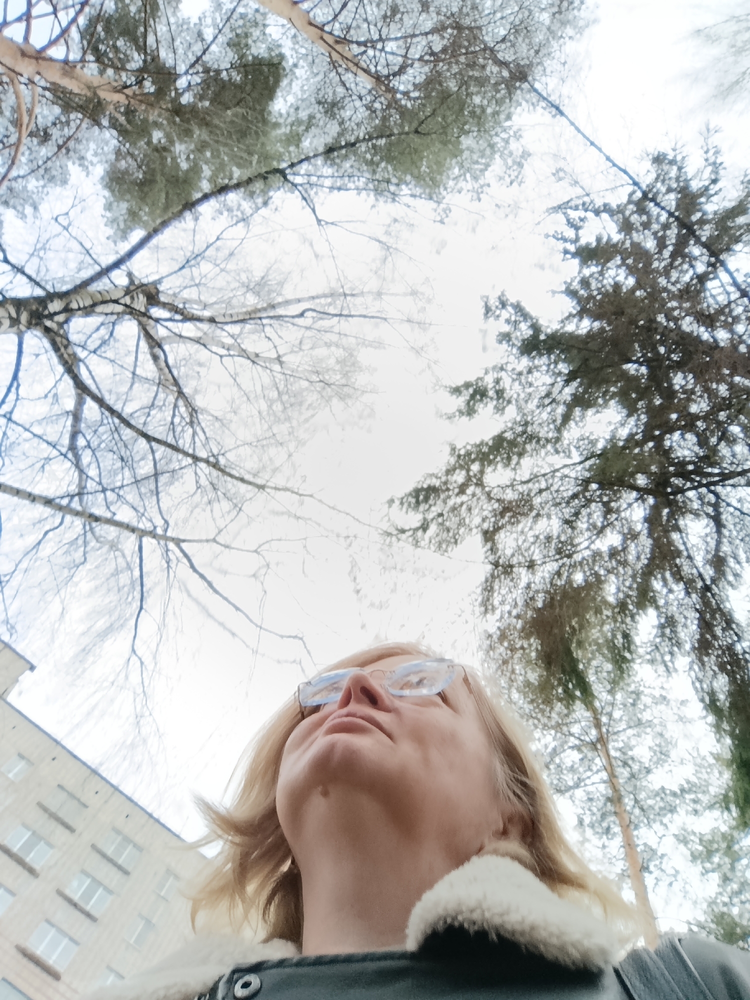 A low-angle, upward-facing photograph captures a woman with blond hair and glasses, looking upwards towards trees against a pale, overcast sky. The trees are a mix of bare branches and dense coniferous foliage. A building is visible to the lower left, with rows of windows. The woman is wearing a jacket with a white, furry collar. The location is Chaikovsky, Russia, based on contextual information not present in the image itself.