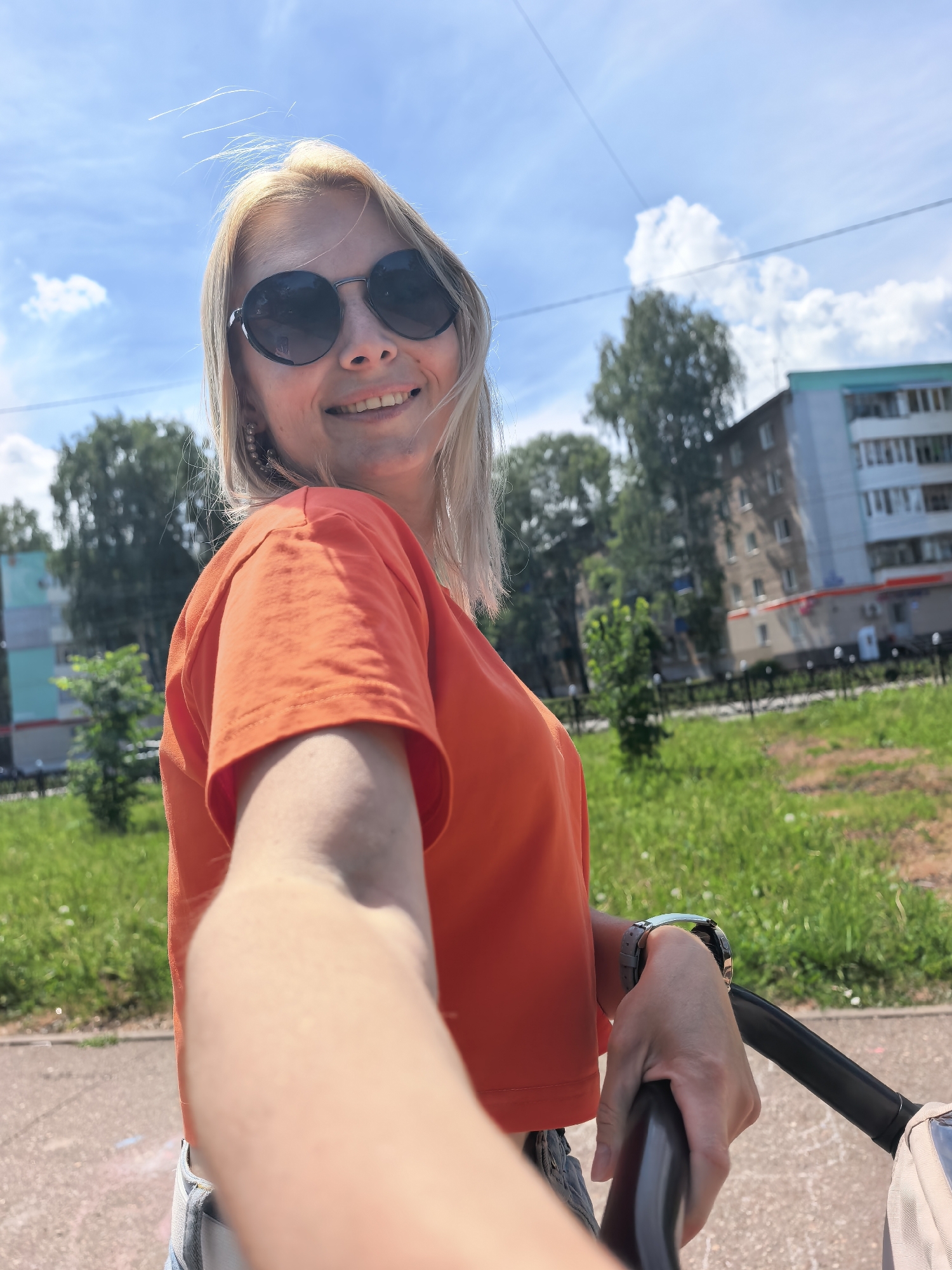 A woman with blonde hair wearing round sunglasses and an orange t-shirt is taking a selfie outdoors. She is smiling and holding the handle of what appears to be a baby stroller. The background includes a grassy area, trees, a sidewalk, and a multi-story building. Based on the metadata, this picture was taken in Sterlitamak, Russia.